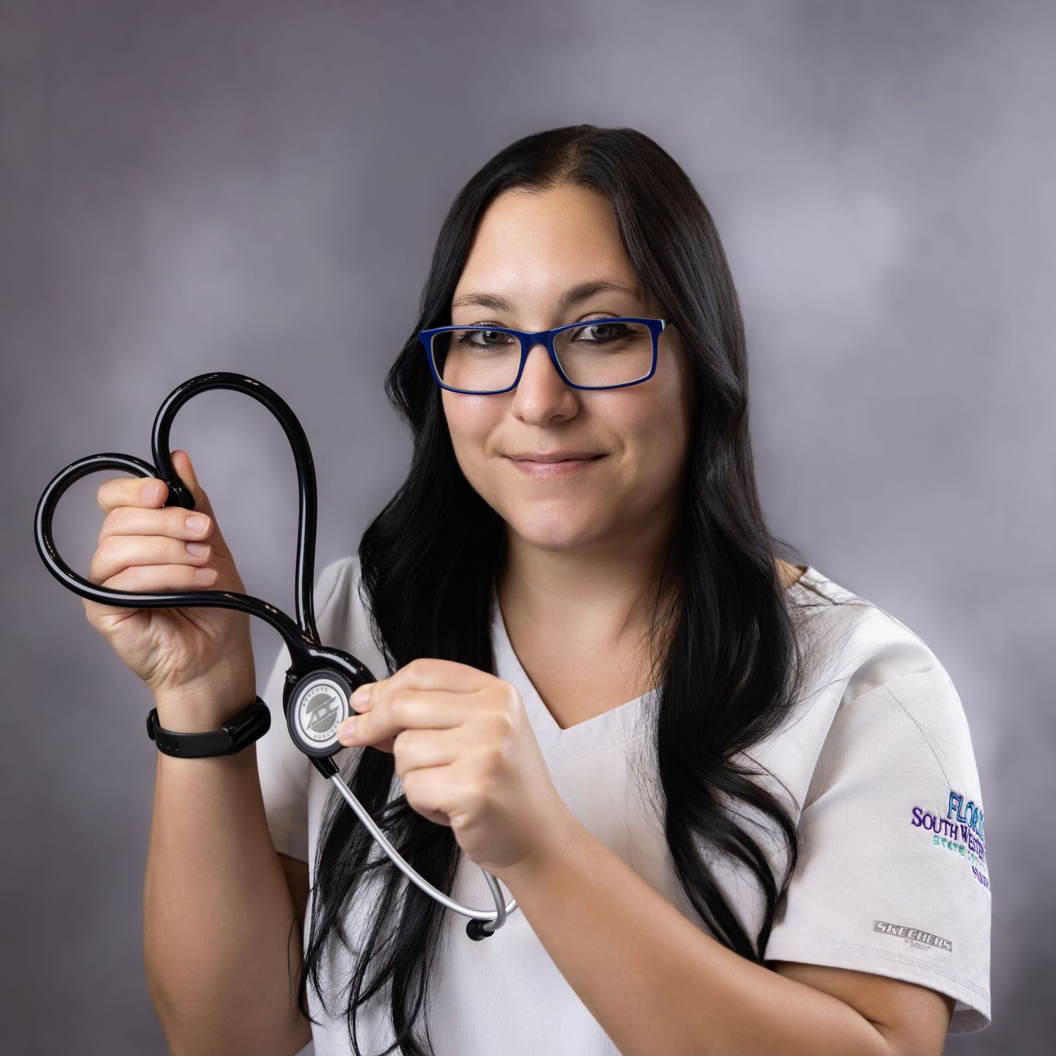 Female nursing student in white scrubs holds stethoscope shaped like heart, wearing glasses, professional studio photo.
