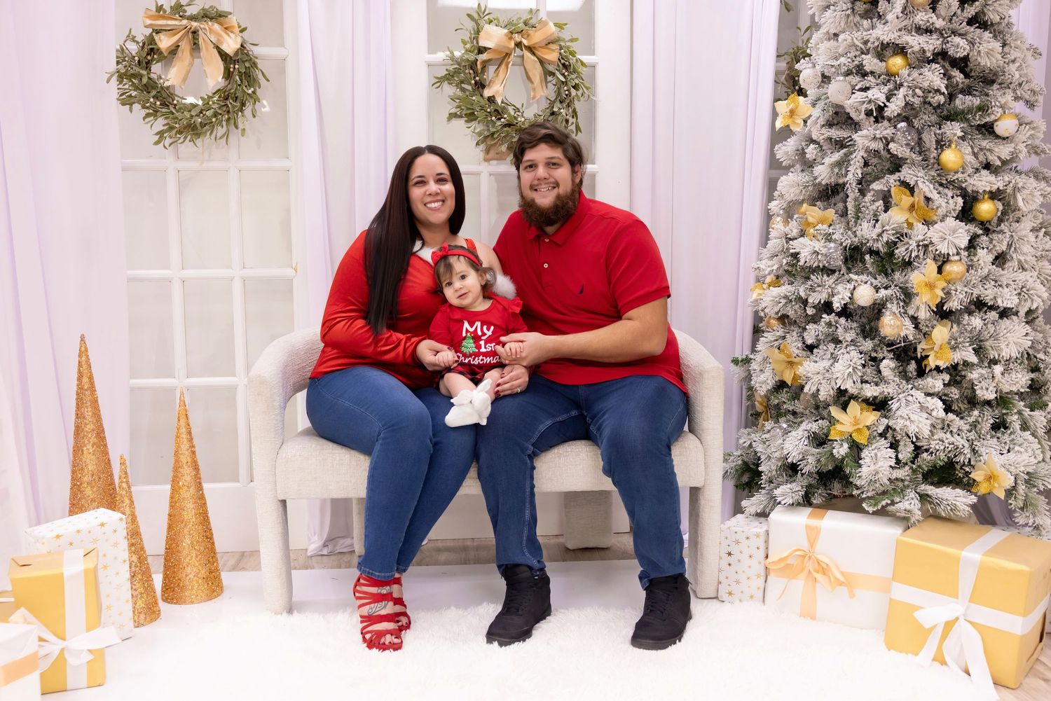 Family of three in red outfits posing for a Christmas portrait beside a decorated tree with gold ornaments and gifts.