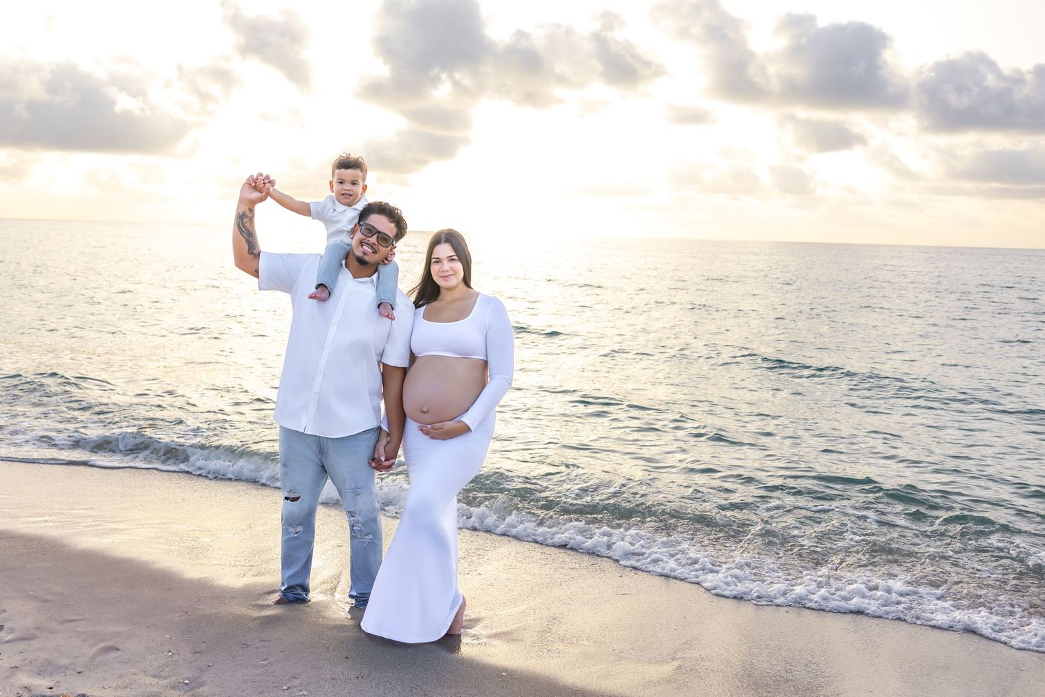 Expecting family poses on beach at sunset, father holds toddler on shoulders beside pregnant mother in white gown.