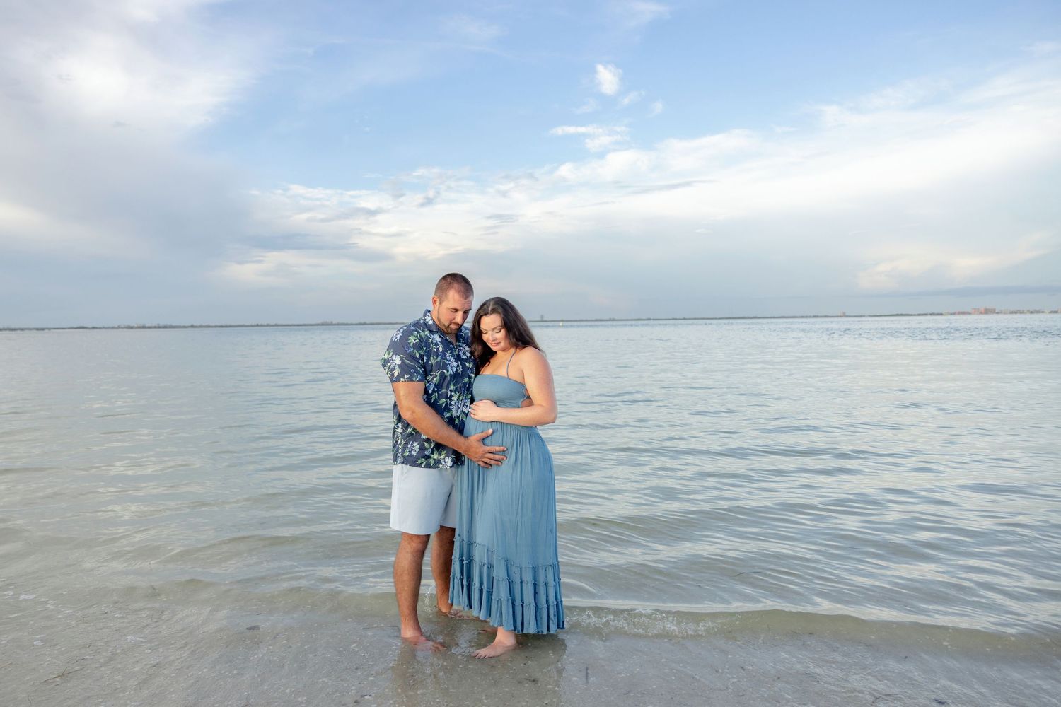 Expecting couple stands barefoot in shallow ocean water at sunset during a beachside maternity photo session, the woman in a blue dress.