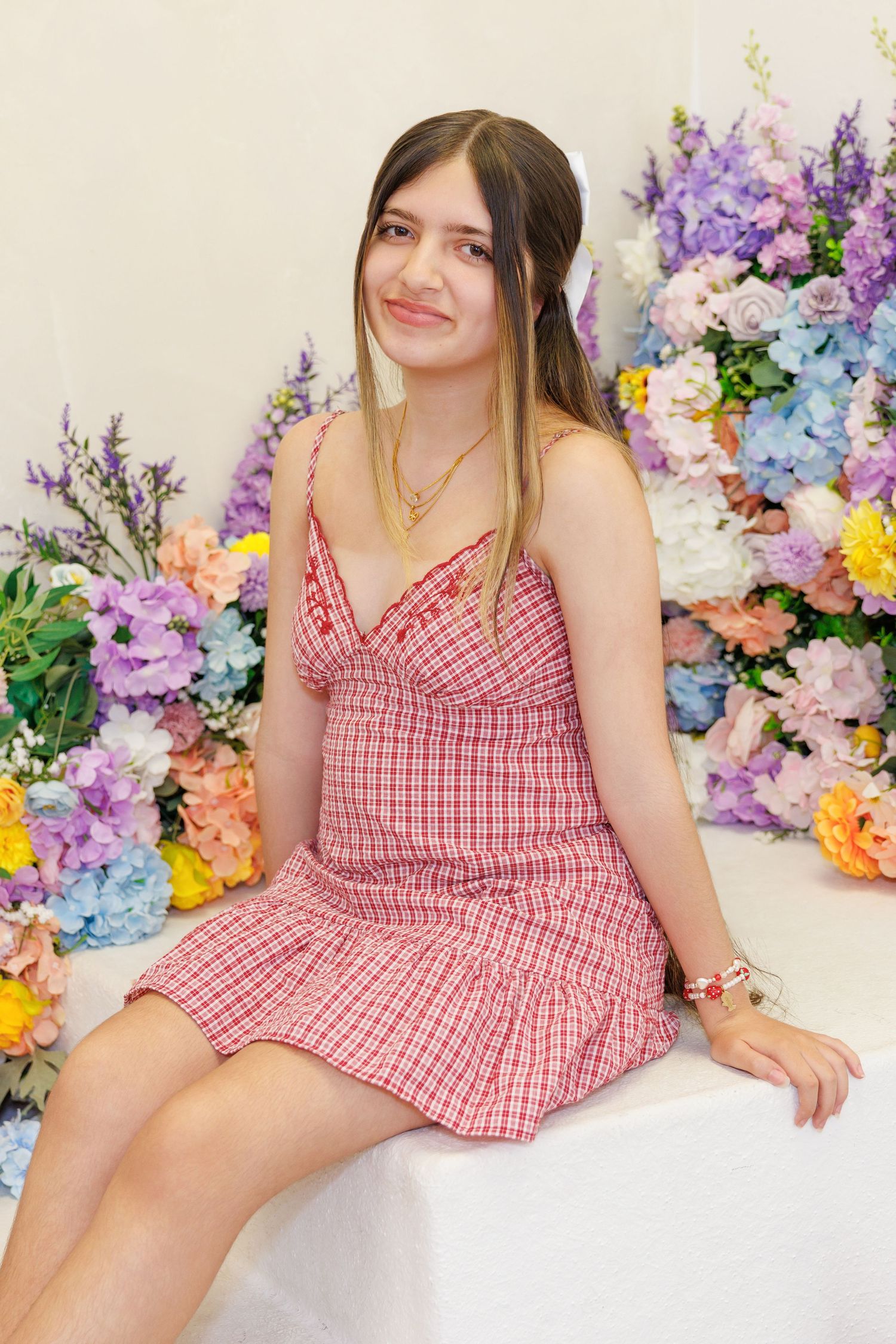 Young woman in red plaid sundress sitting in front of colorful floral arrangement backdrop, smiling softly.