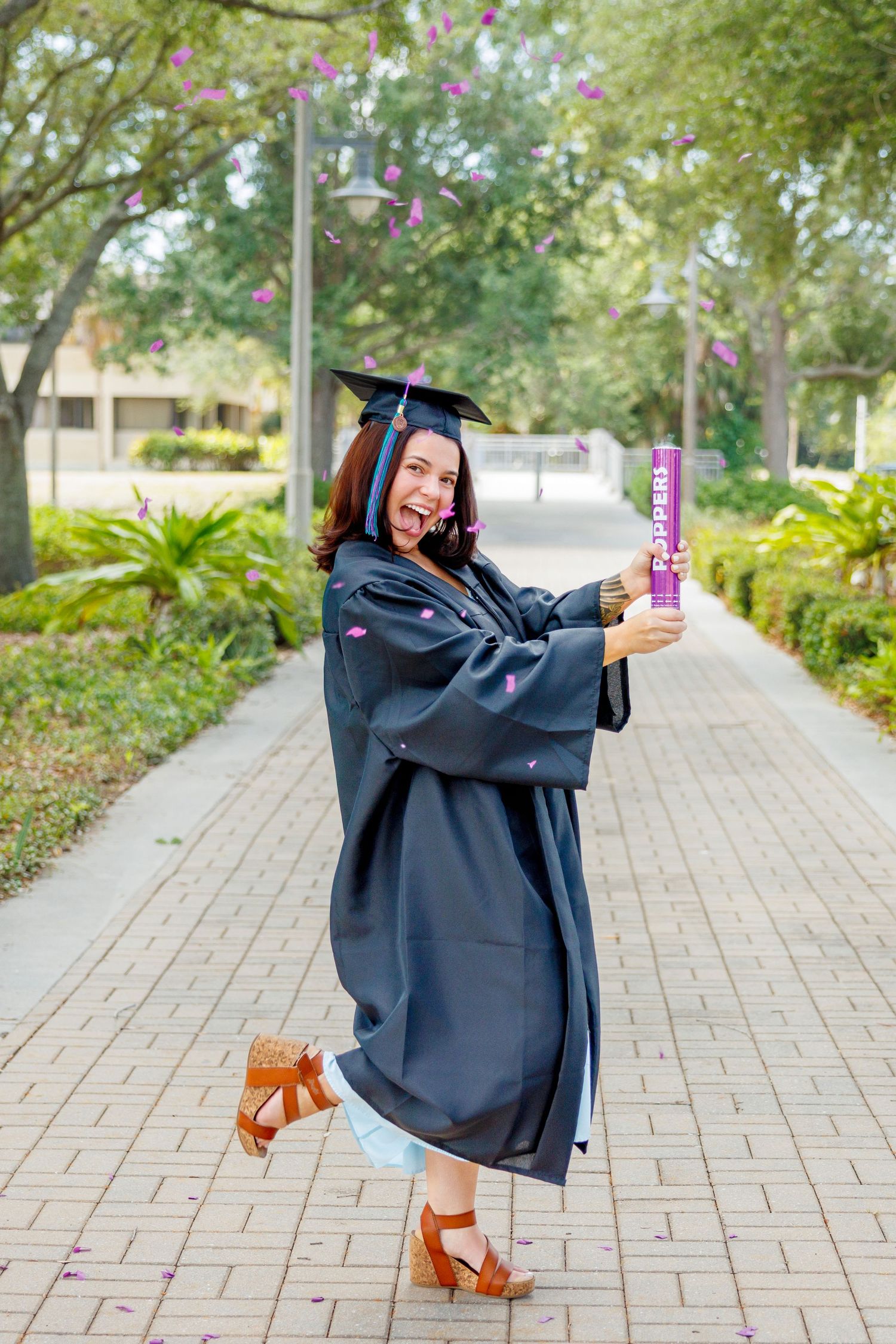 Smiling graduate in a black cap and gown poses on a brick path, firing a purple confetti popper while pink petals float through the air.