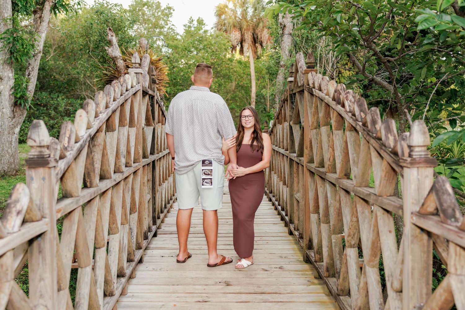 Couple standing arm in arm on a rustic wooden bridge with carved railings, surrounded by lush tropical greenery and palm trees.