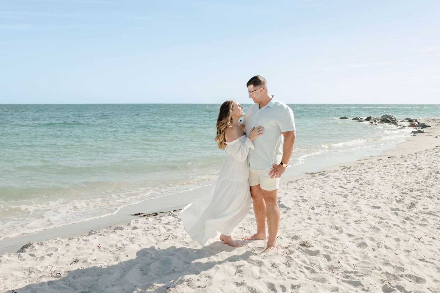 Couple in white outfits embracing barefoot on a sandy beach with calm turquoise water and a clear blue sky.