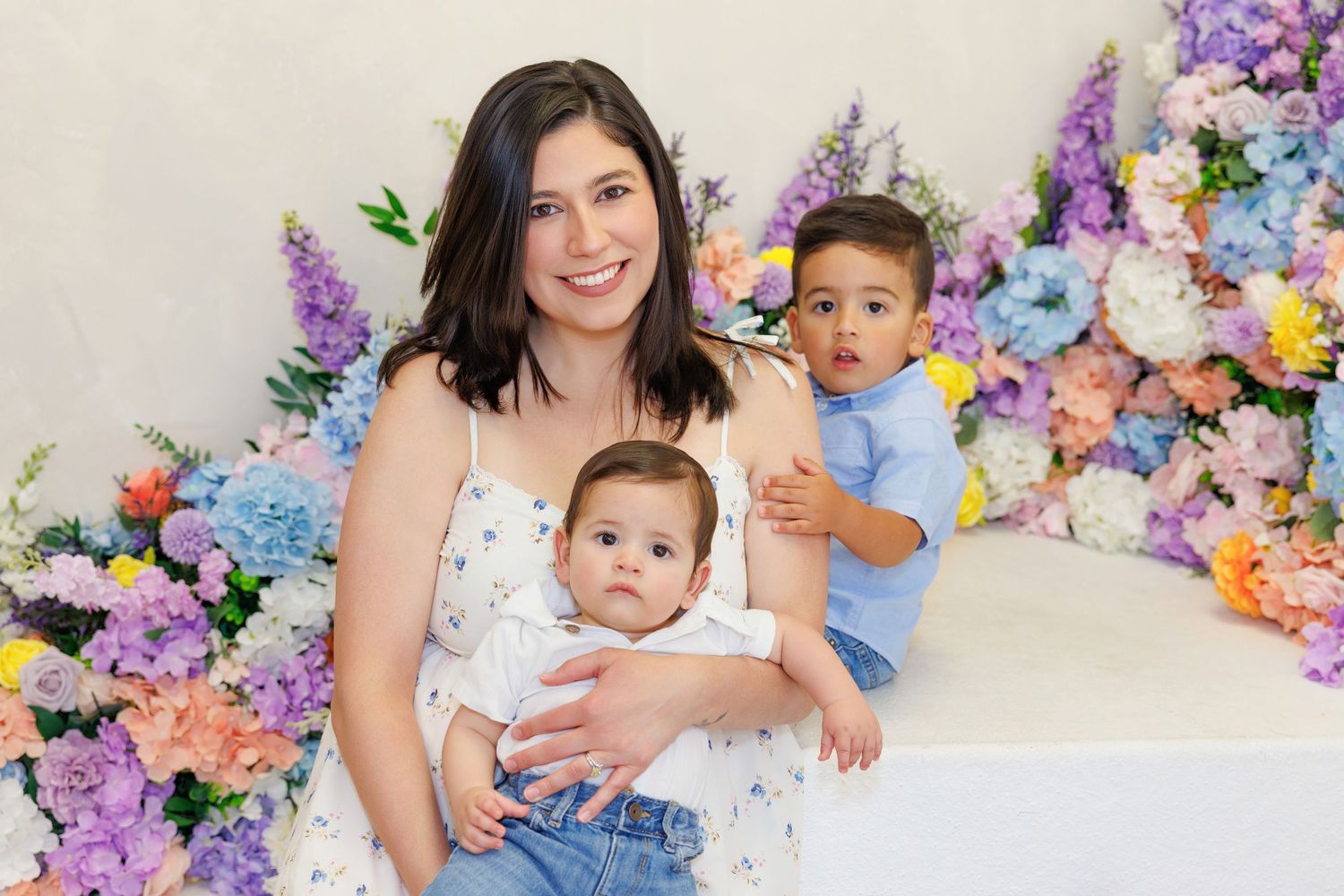 A smiling mother poses with her baby and toddler son in front of a colorful floral backdrop.