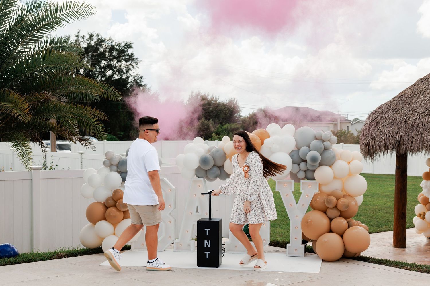 Couple walking outdoors at a gender reveal party with pink smoke, balloon garlands, a TNT box, and a thatched tiki hut.