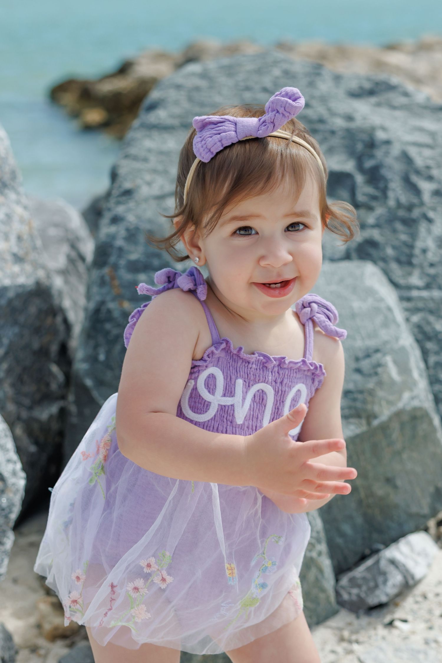 Smiling toddler girl in purple floral dress and bow headband posing near rocky beach shore.