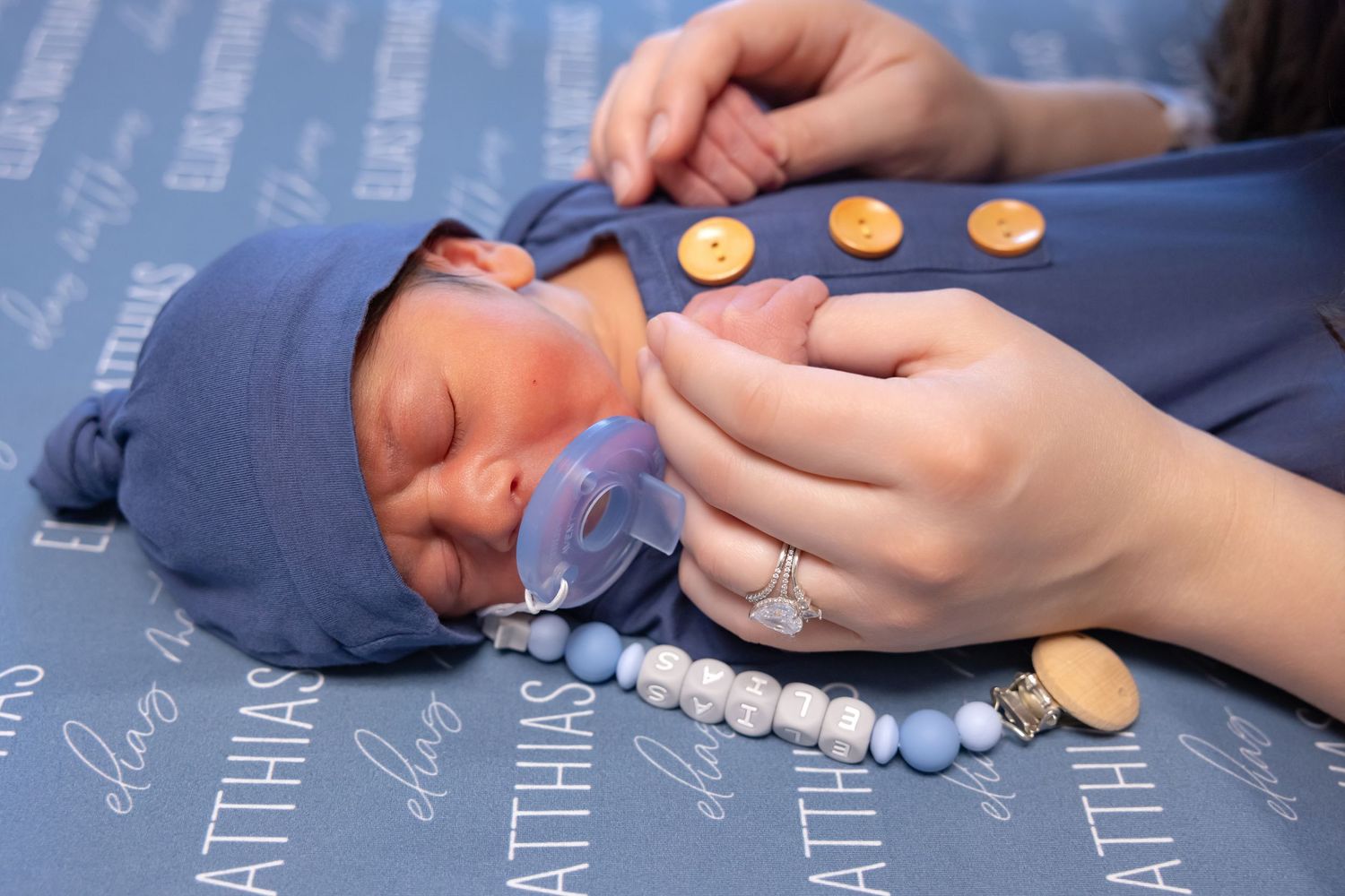 Sleeping newborn baby in blue outfit with pacifier and personalized beaded clip on blue blanket with names.