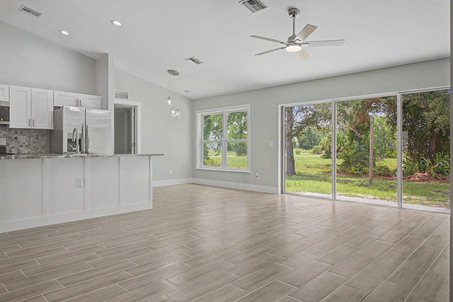 Bright open-concept living area with vaulted ceiling, ceiling fan, wood-look tile floors, kitchen and sliding glass doors to a green yard.