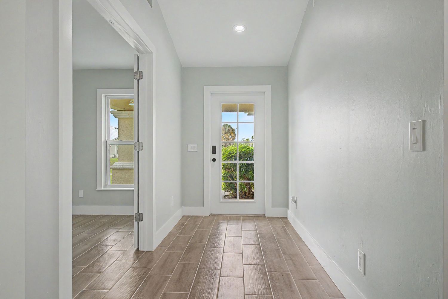 Bright hallway with wood-look plank tile flooring, gray walls, white trim, and a glass-paneled door leading to a sunny outdoor view.