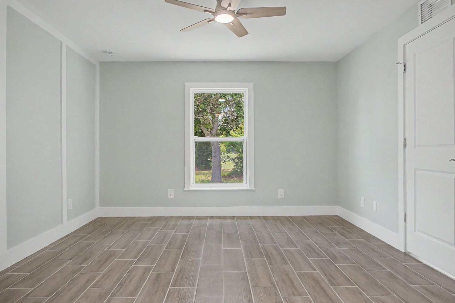Empty bedroom with light sage green walls, wood-look plank tile flooring, a ceiling fan, and a single window overlooking green trees.