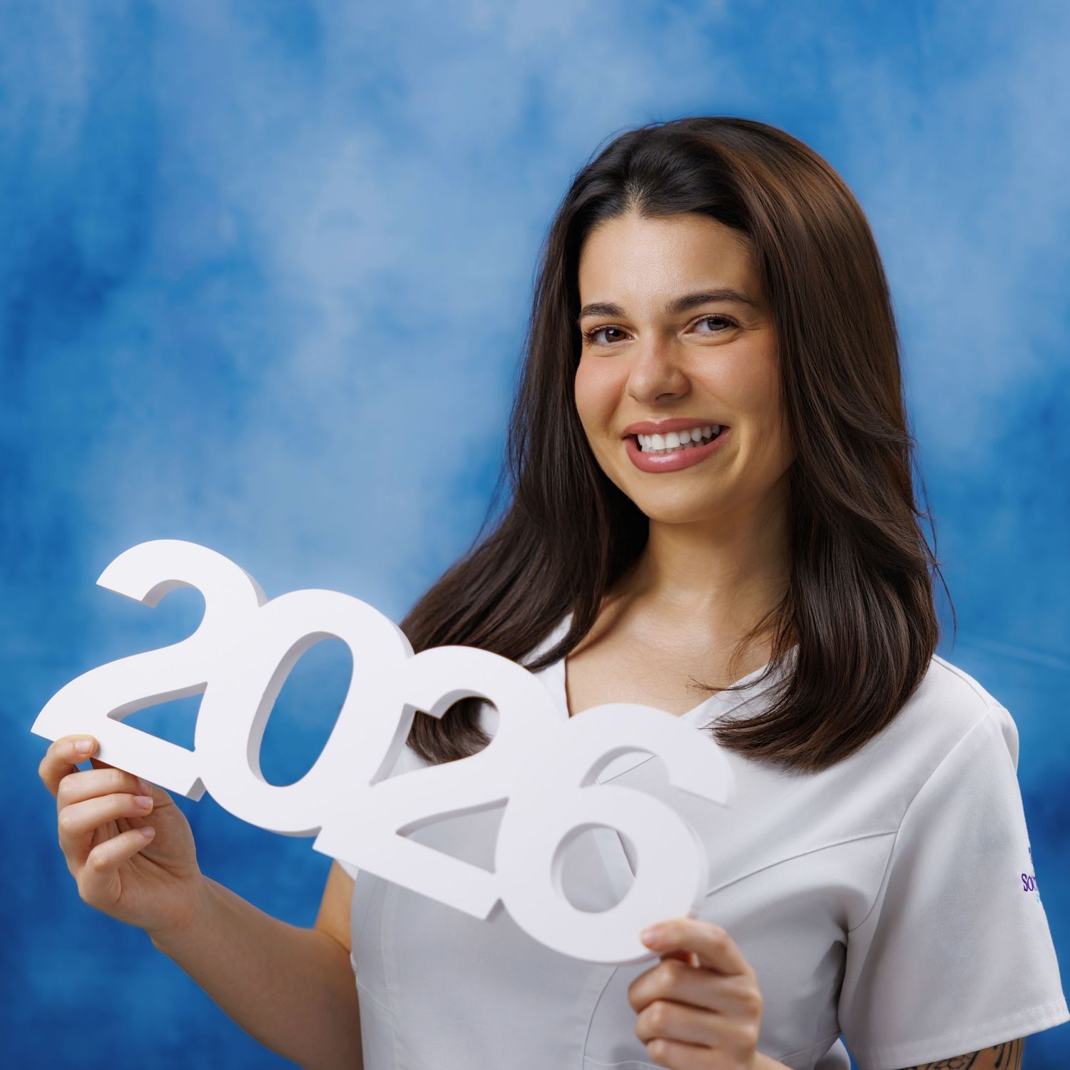 Smiling young woman holding a white 2026 sign against a blue background, perfect for graduation announcements.