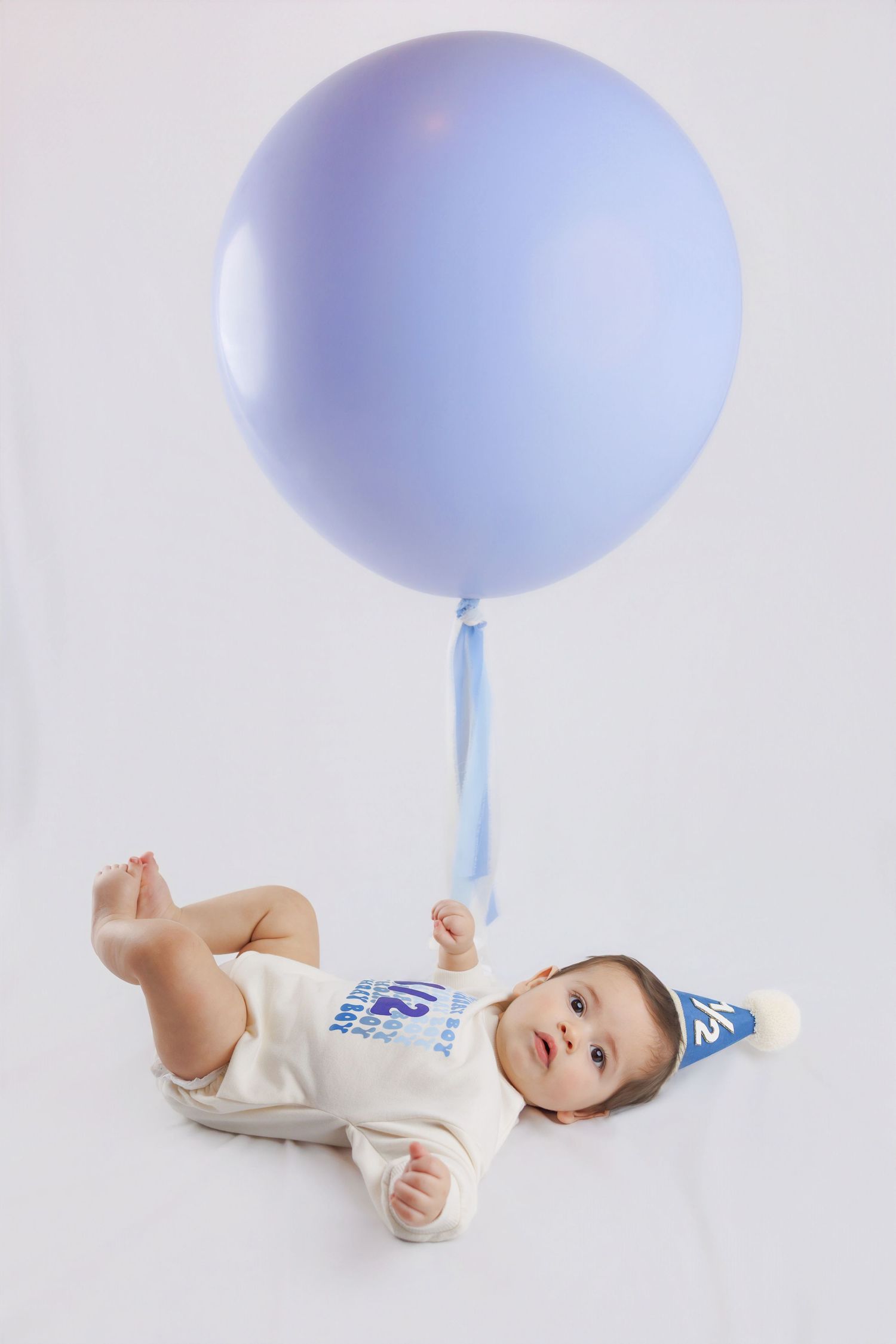 Cute baby lying down holding a large lavender balloon with a blue half birthday hat on a white backdrop.