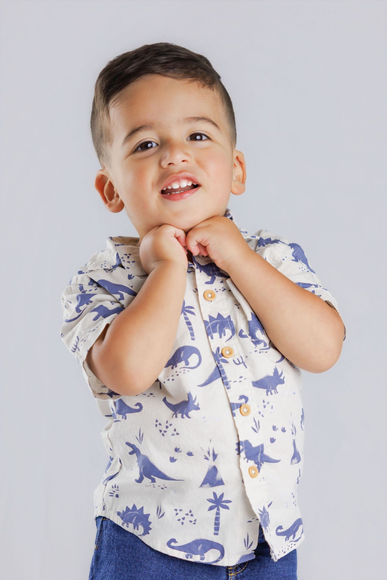 Happy toddler boy in a dinosaur print shirt posing with hands under chin against a gray background.