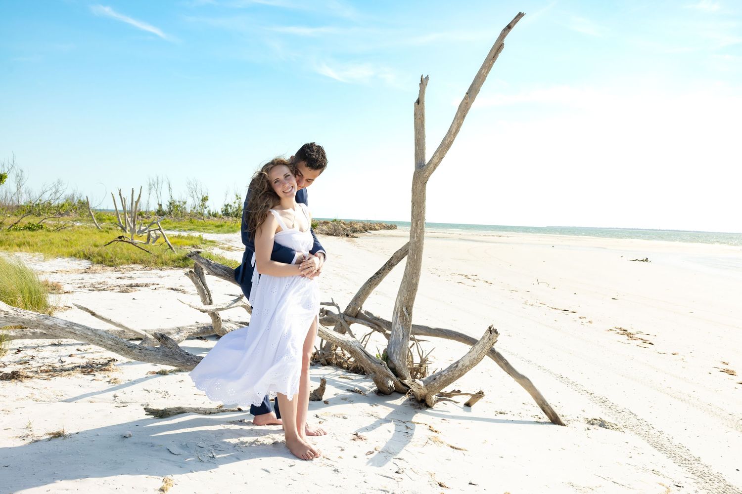 Couple in wedding attire embracing on a white sandy beach beside driftwood under a clear blue sky.