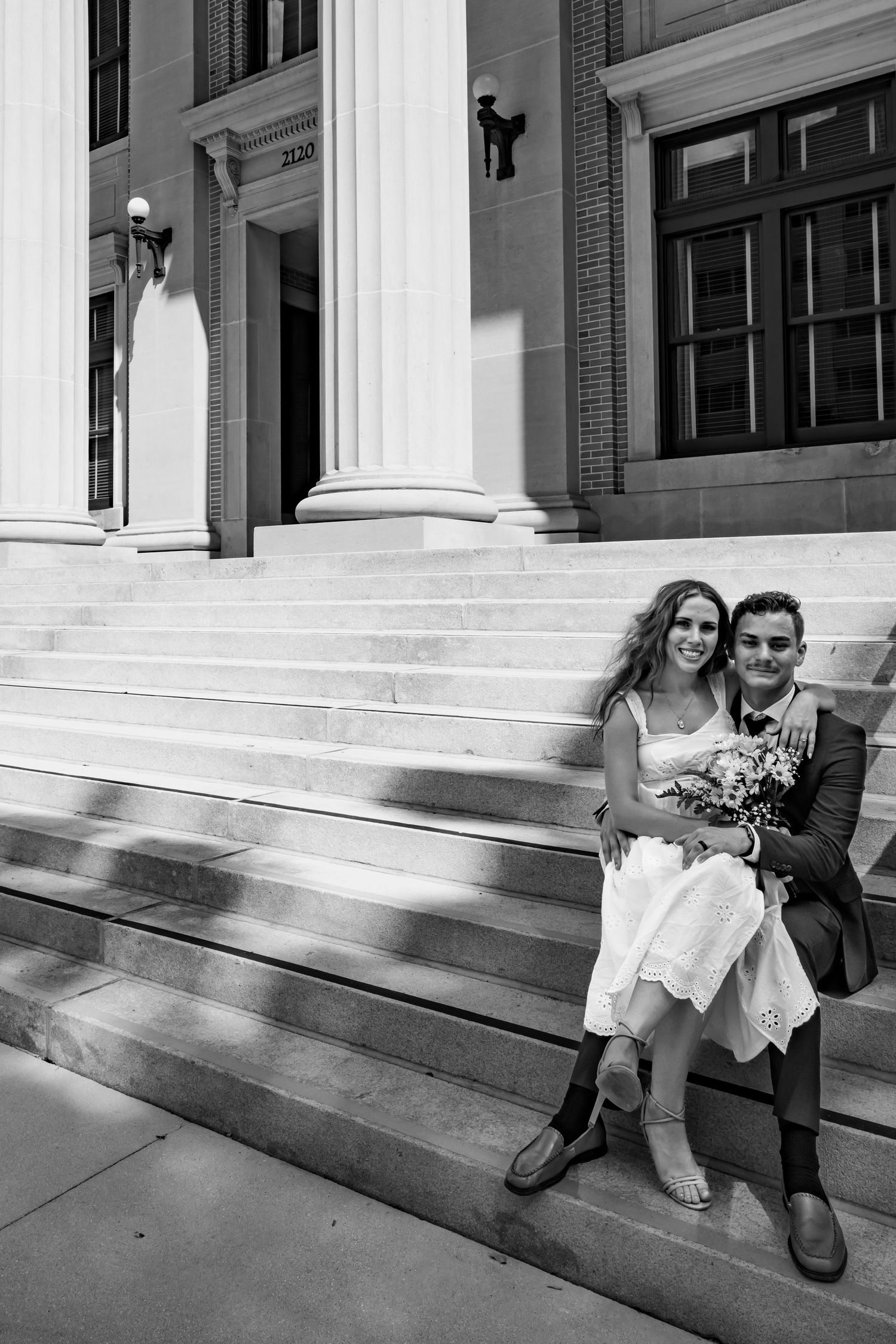 Black and white photo of a couple in wedding attire sitting on steps of a classic building with columns.