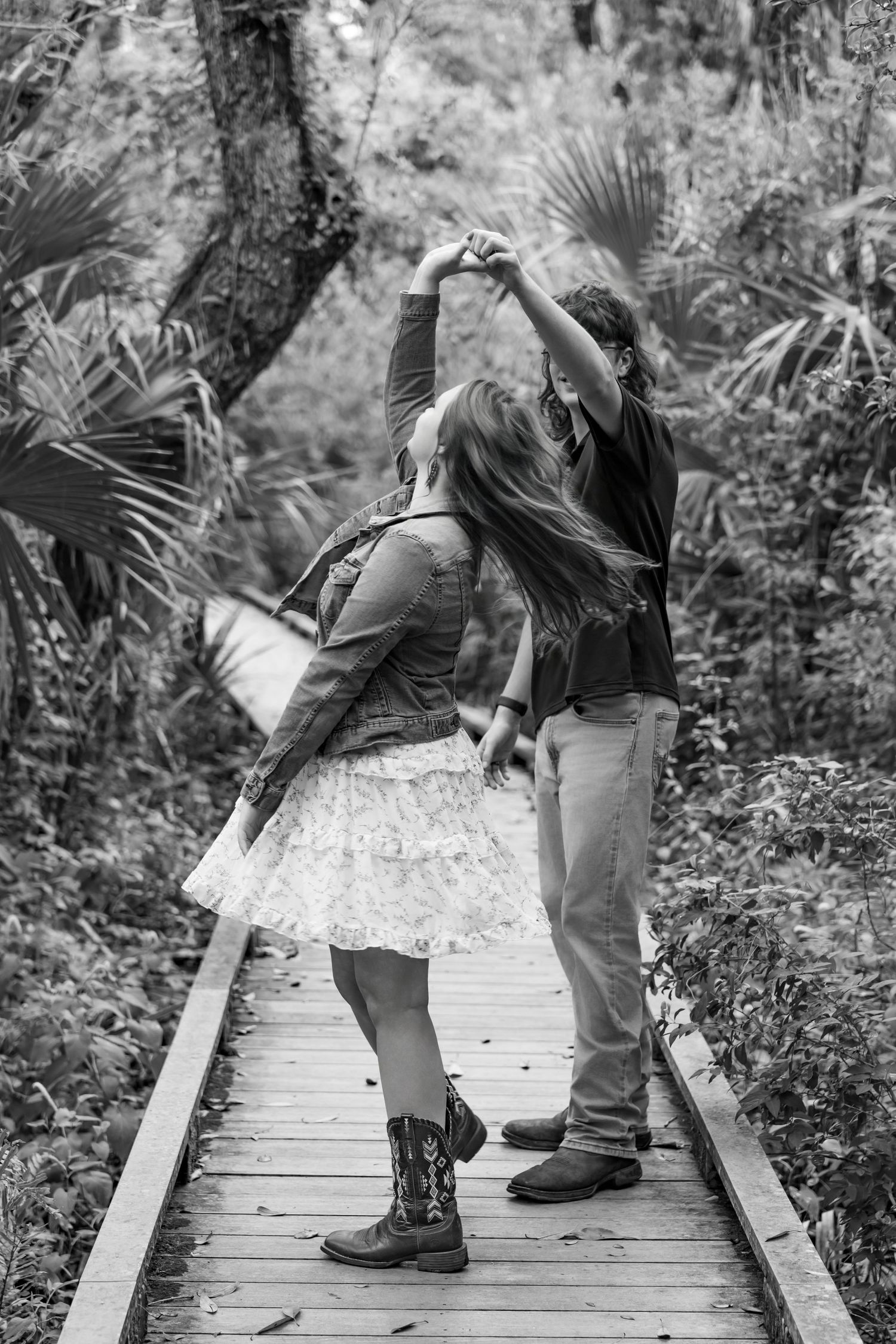 Couple dancing on a wooden boardwalk path surrounded by lush greenery in a black and white photo.