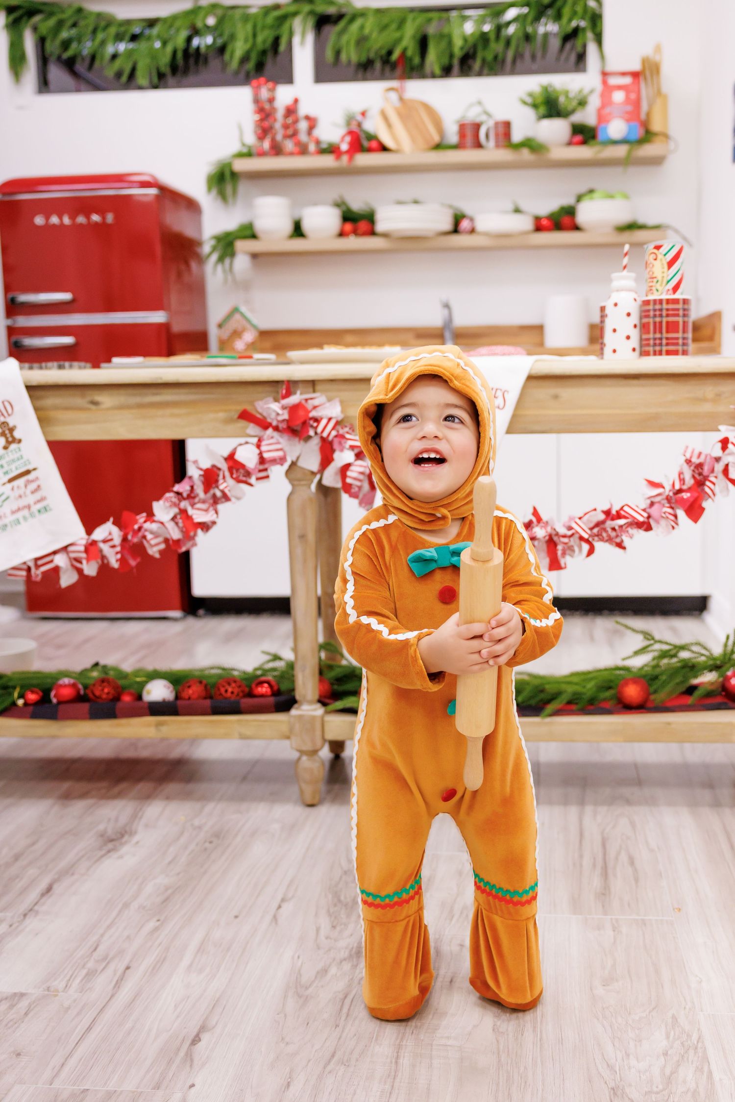 Toddler in gingerbread man costume holding rolling pin in Christmas decorated kitchen.