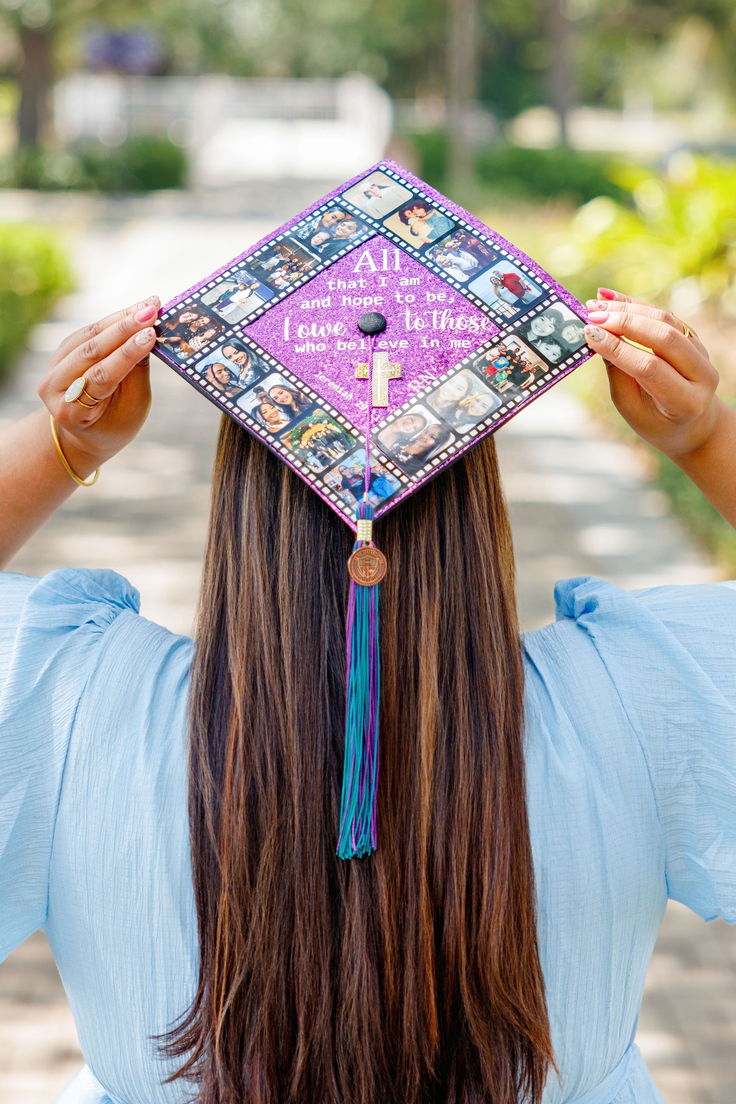 Graduate with long brown hair holds a purple decorated graduation cap with photo collage, cross, and teal tassel outdoors.