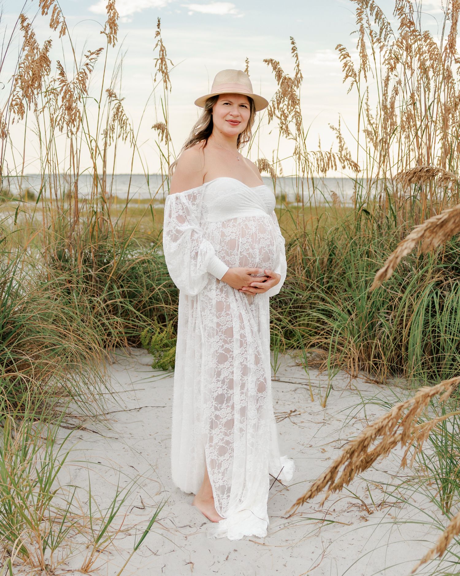 Pregnant woman in a white lace maternity gown and straw hat cradles her baby bump on a sandy beach dune surrounded by tall sea oats.