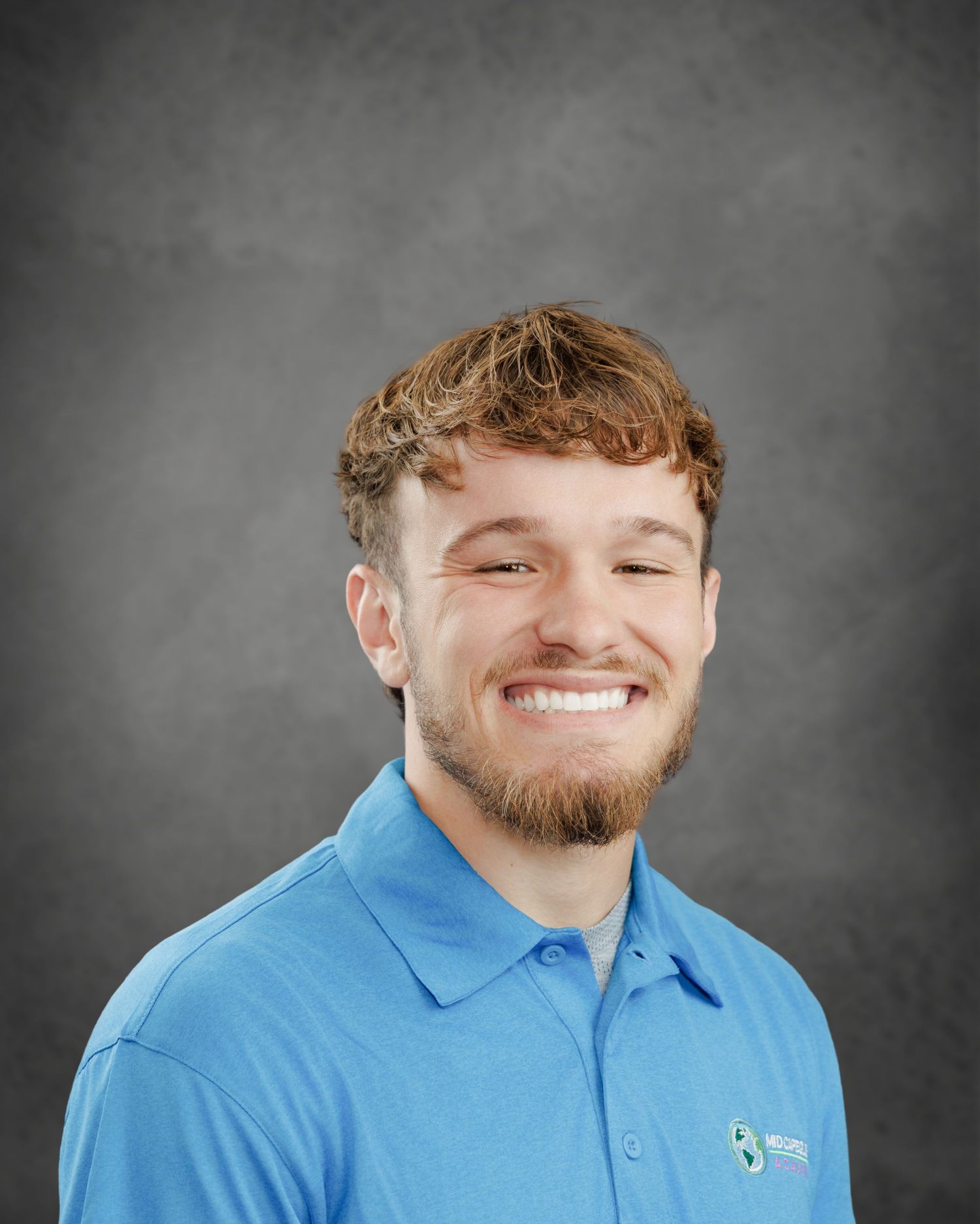 Smiling young man with tousled red-brown hair and a beard wearing a blue polo shirt with a small logo against a gray backdrop.