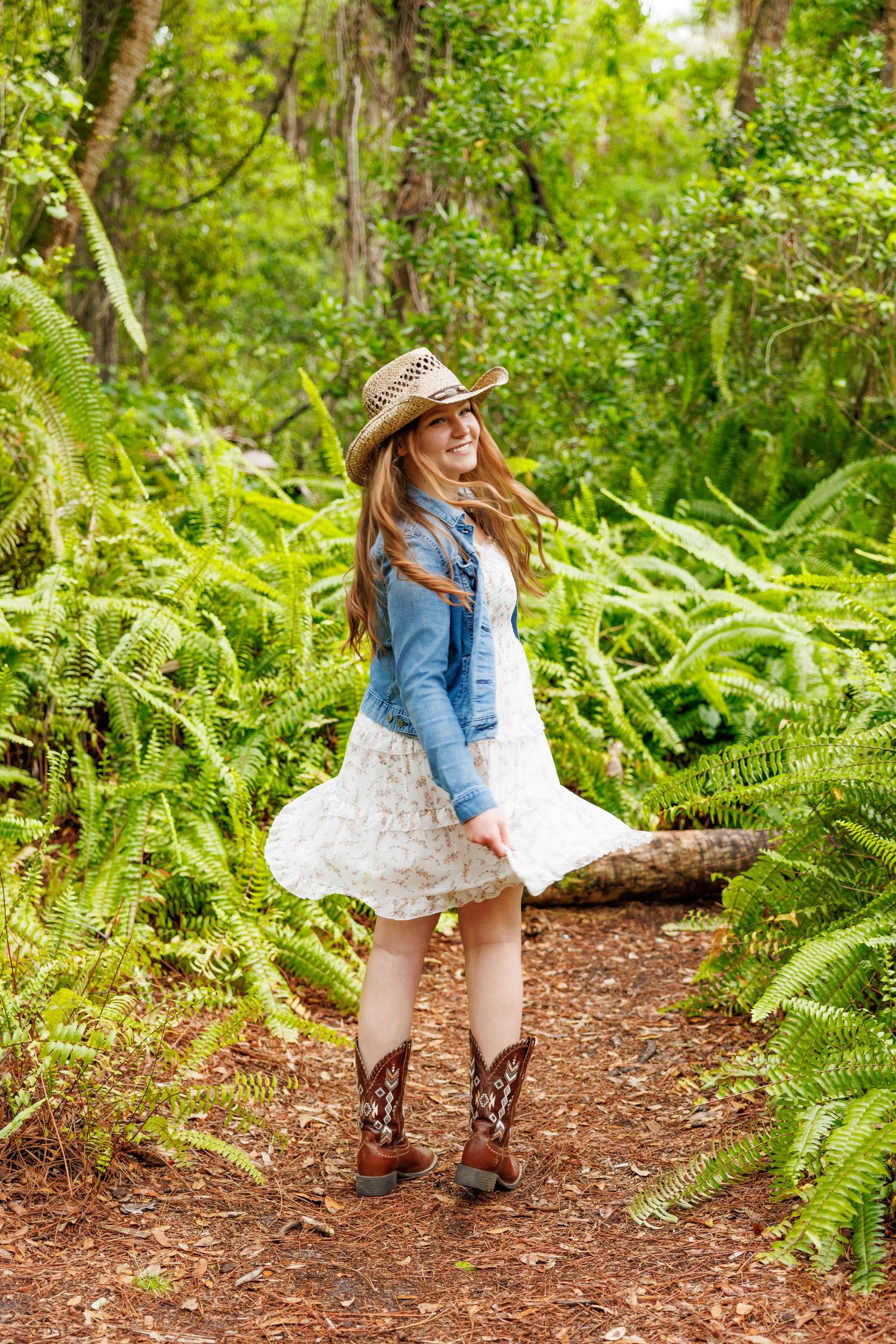 Young woman in cowboy hat, denim jacket, white skirt, and boots twirling on a forest path surrounded by ferns.