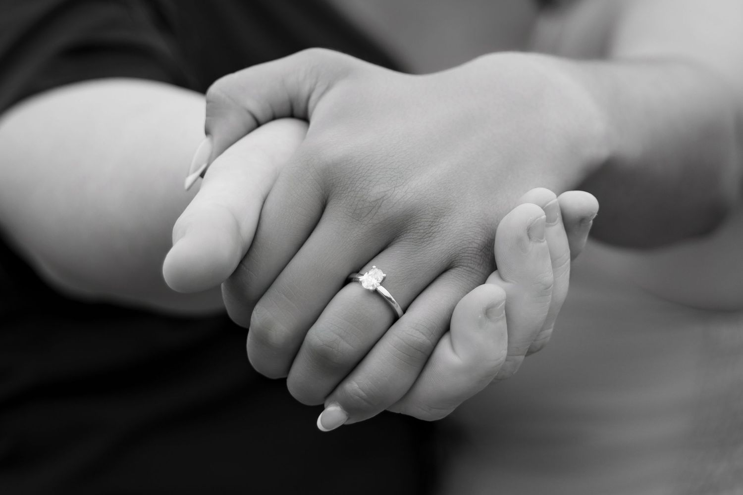 Black and white photo of two hands holding, woman wearing a diamond engagement ring, romantic couple moment.
