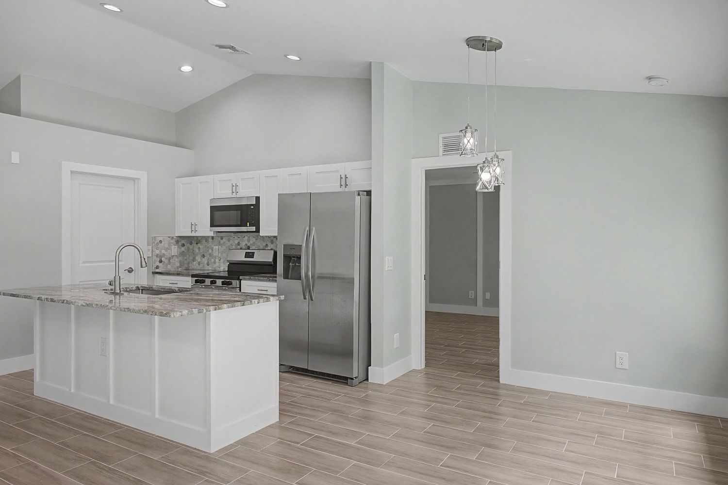 Open-concept kitchen with white cabinets, granite island, stainless steel appliances, pendant lighting and wood-look tile flooring.