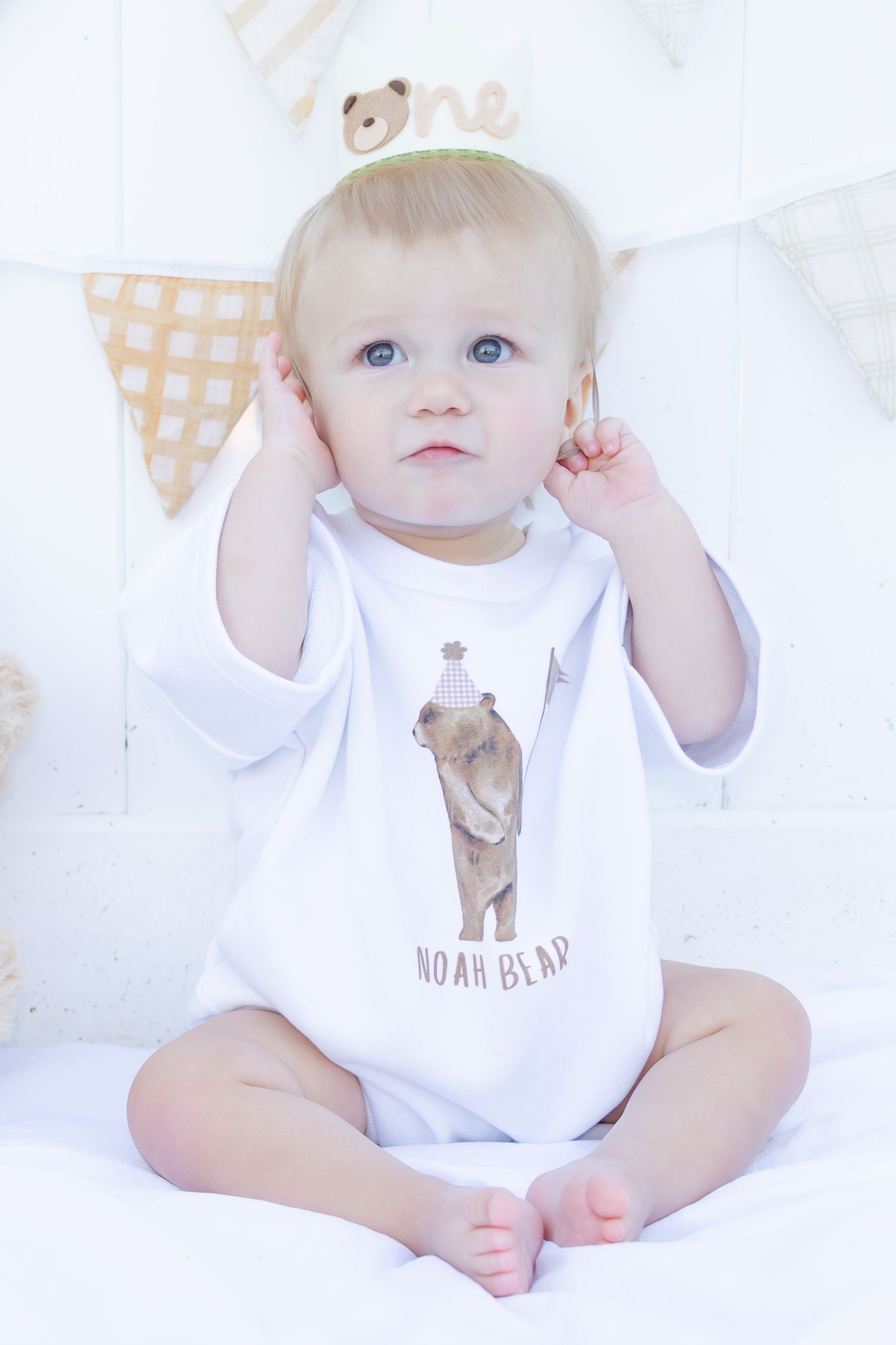 Baby in white Noah Bear onesie with party hat sits cross-legged against festive bunting backdrop.
