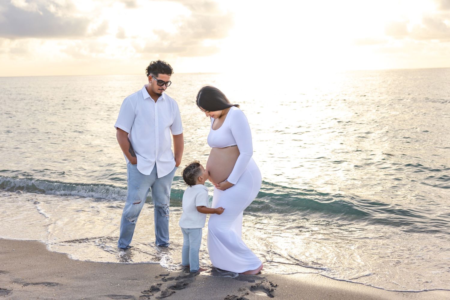 Pregnant woman in white dress with husband and child on beach at sunset.