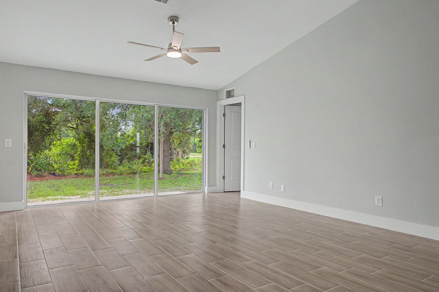 Empty living room with vaulted ceiling, ceiling fan, wood-look tile flooring, and sliding glass doors overlooking a green backyard.