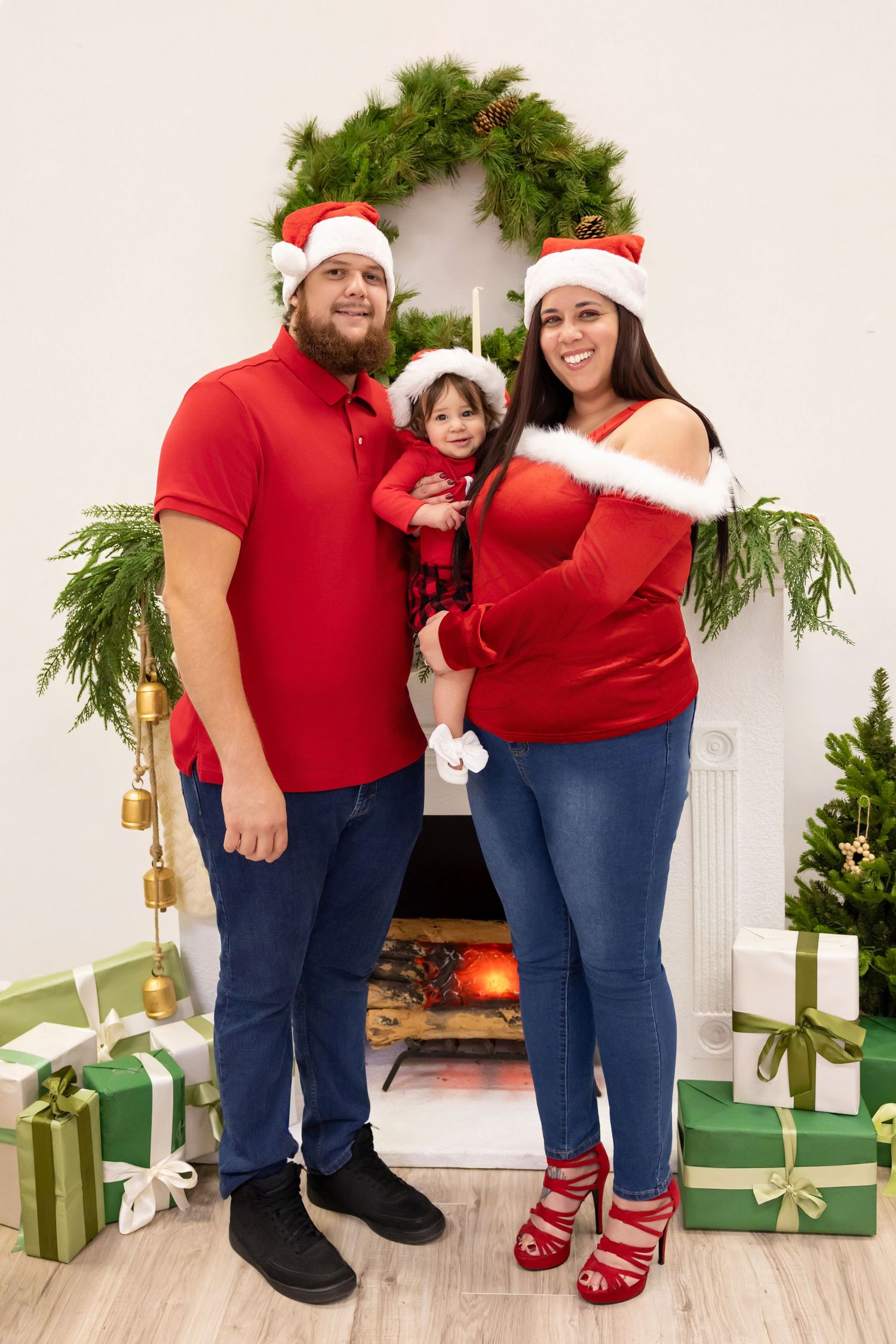 Family of three in matching red Santa hats pose by a decorated fireplace with Christmas gifts and greenery.