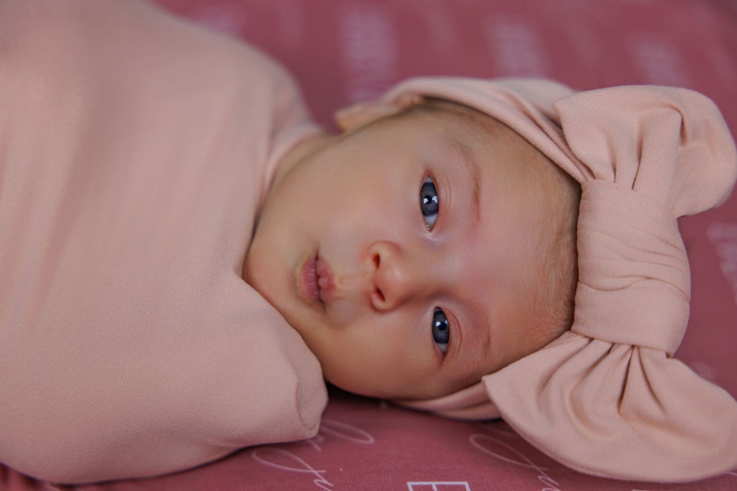 Newborn baby with blue eyes wearing a blush pink bow headband and swaddled in a soft pink wrap, lying on a pink patterned blanket.
