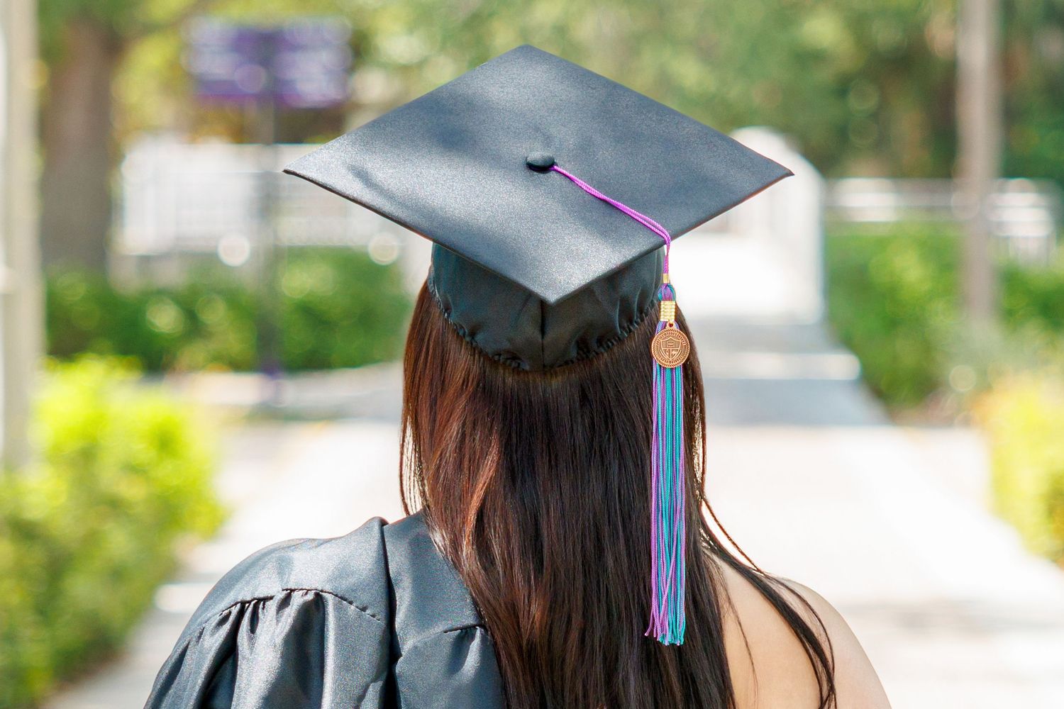 Rear view of a graduate with long brown hair wearing a black cap and gown with a purple and blue tassel on an outdoor campus path.