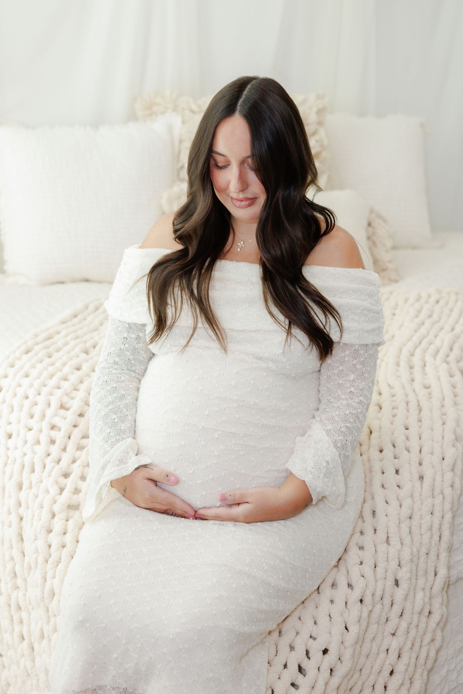 Pregnant woman in a white off-the-shoulder lace maternity gown cradles her belly while seated on a bed with cream chunky knit blanket.