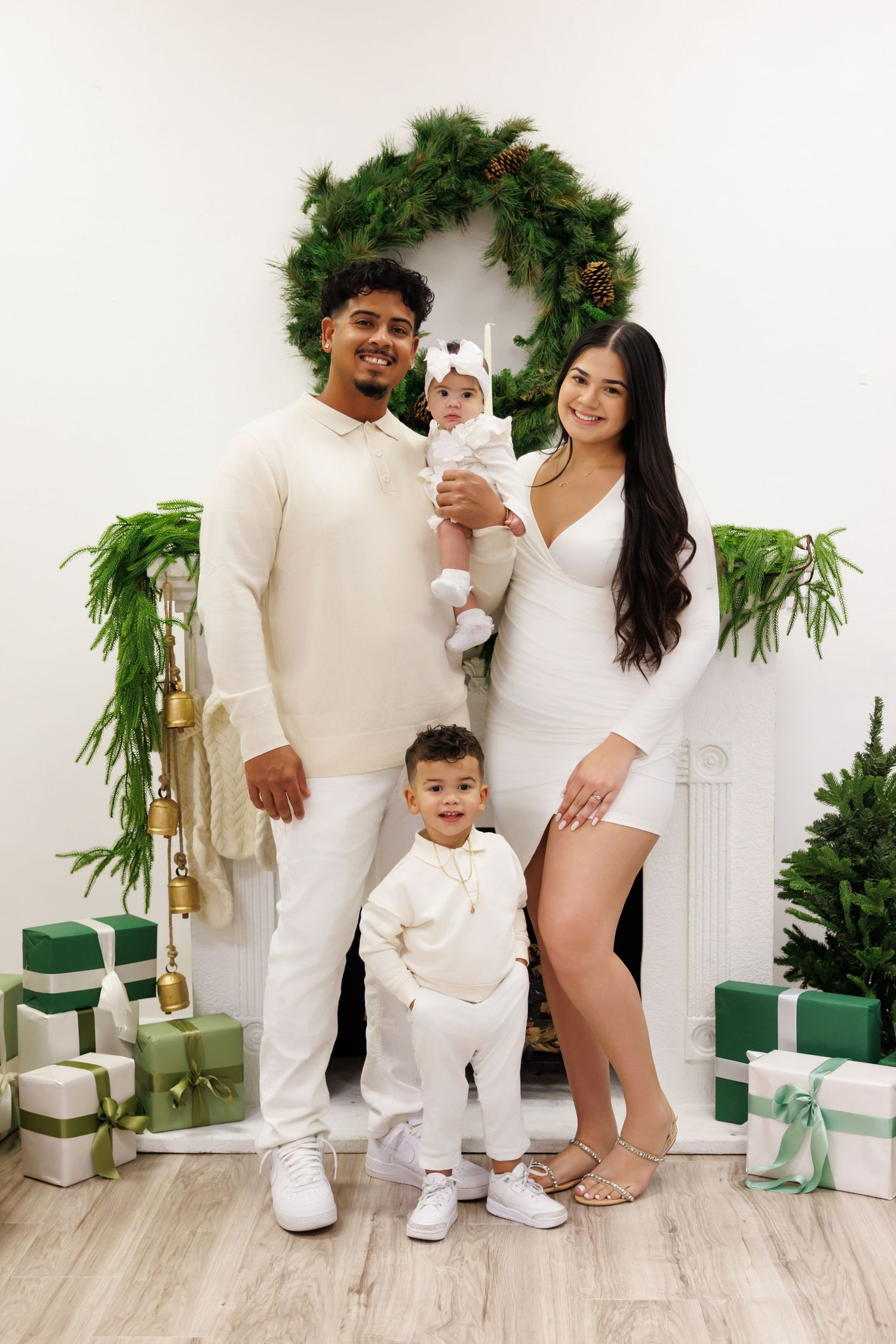 Family of four in white outfits poses by decorated fireplace with Christmas wreath and green wrapped gifts.