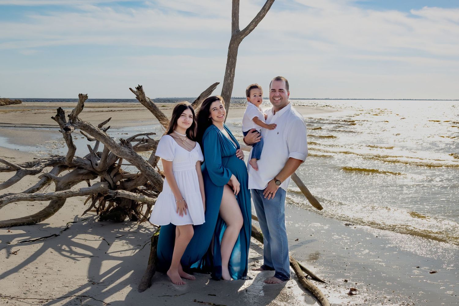 Family of four posing on a sandy beach near driftwood, with calm ocean waters in the background on a sunny day.