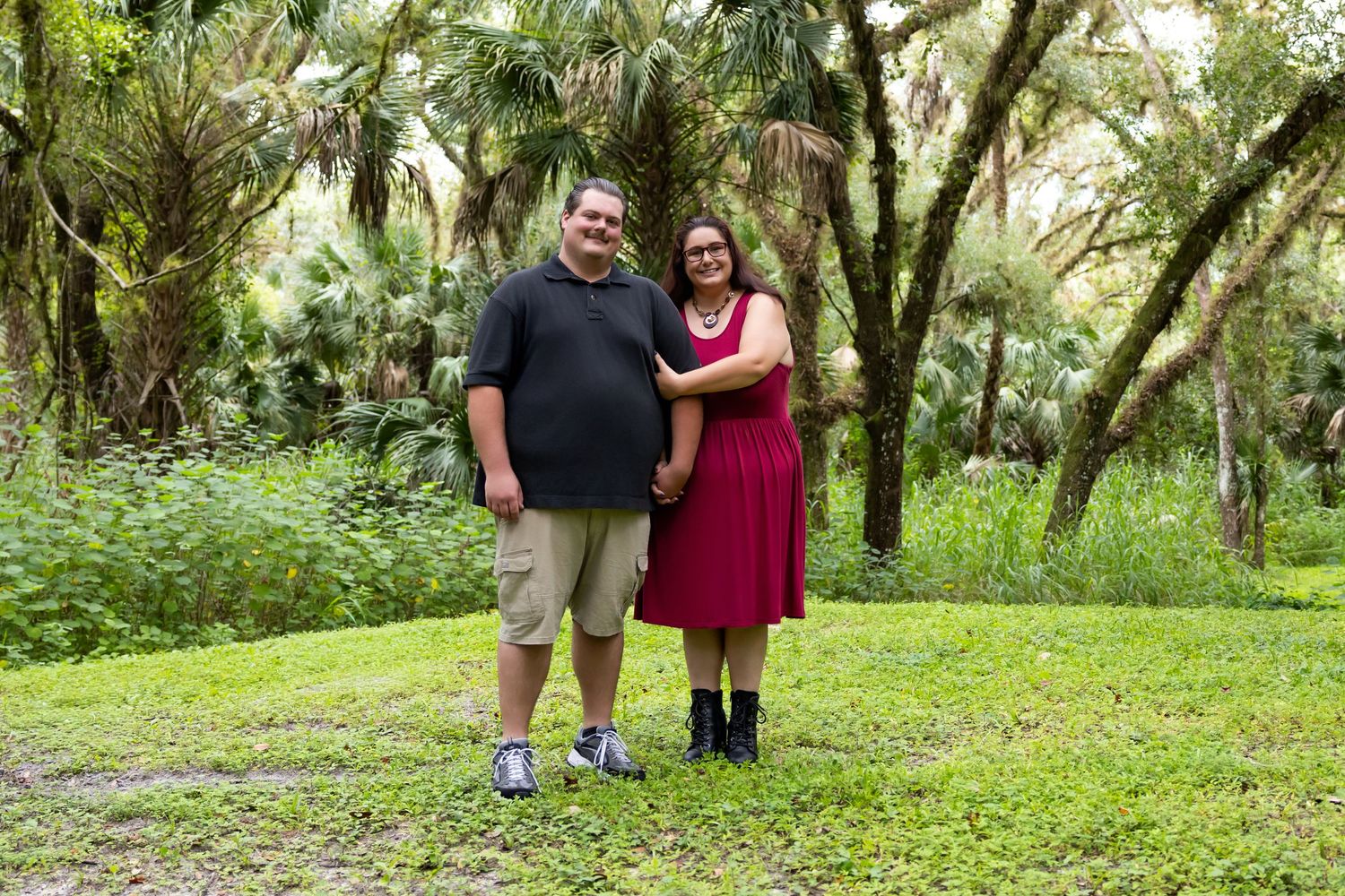 Couple smiling and holding hands in a lush green park surrounded by palm trees and moss-covered oaks.