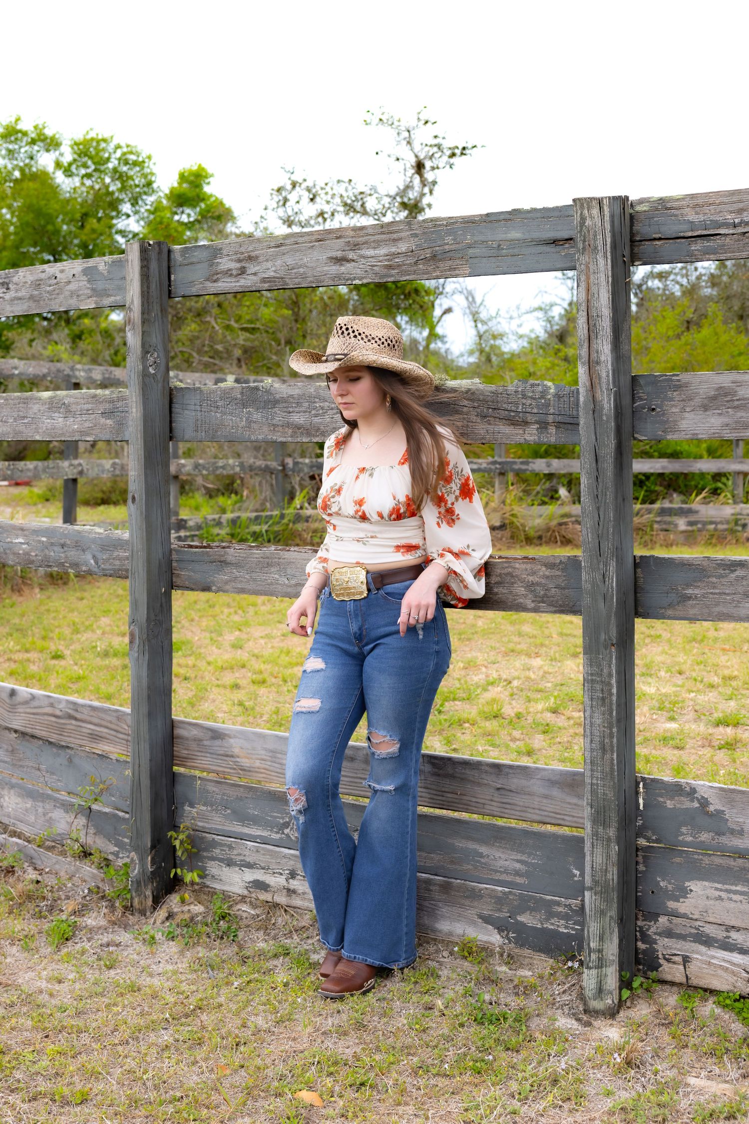 Young woman in cowboy hat, floral top, ripped flare jeans, leaning against rustic wooden fence on a farm.