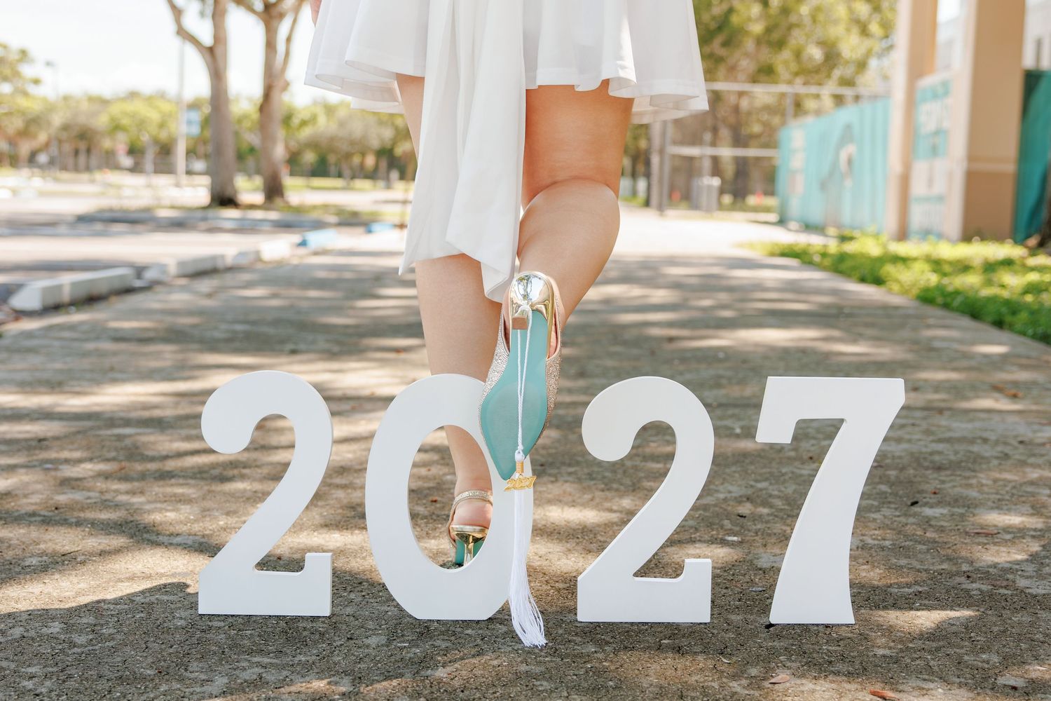 Graduate in a white dress and sandals poses outdoors with large white numbers spelling 2027, holding a teal graduation cap by its tassel.