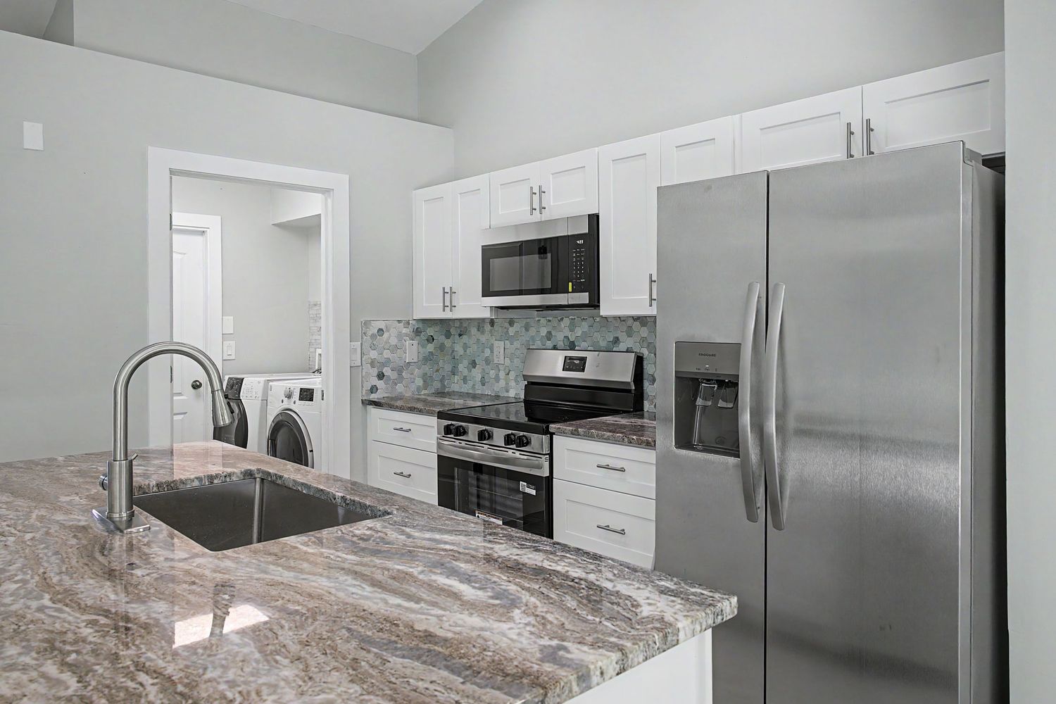 Modern kitchen with white cabinets, stainless steel appliances, mosaic tile backsplash, and a granite island with an undermount sink.