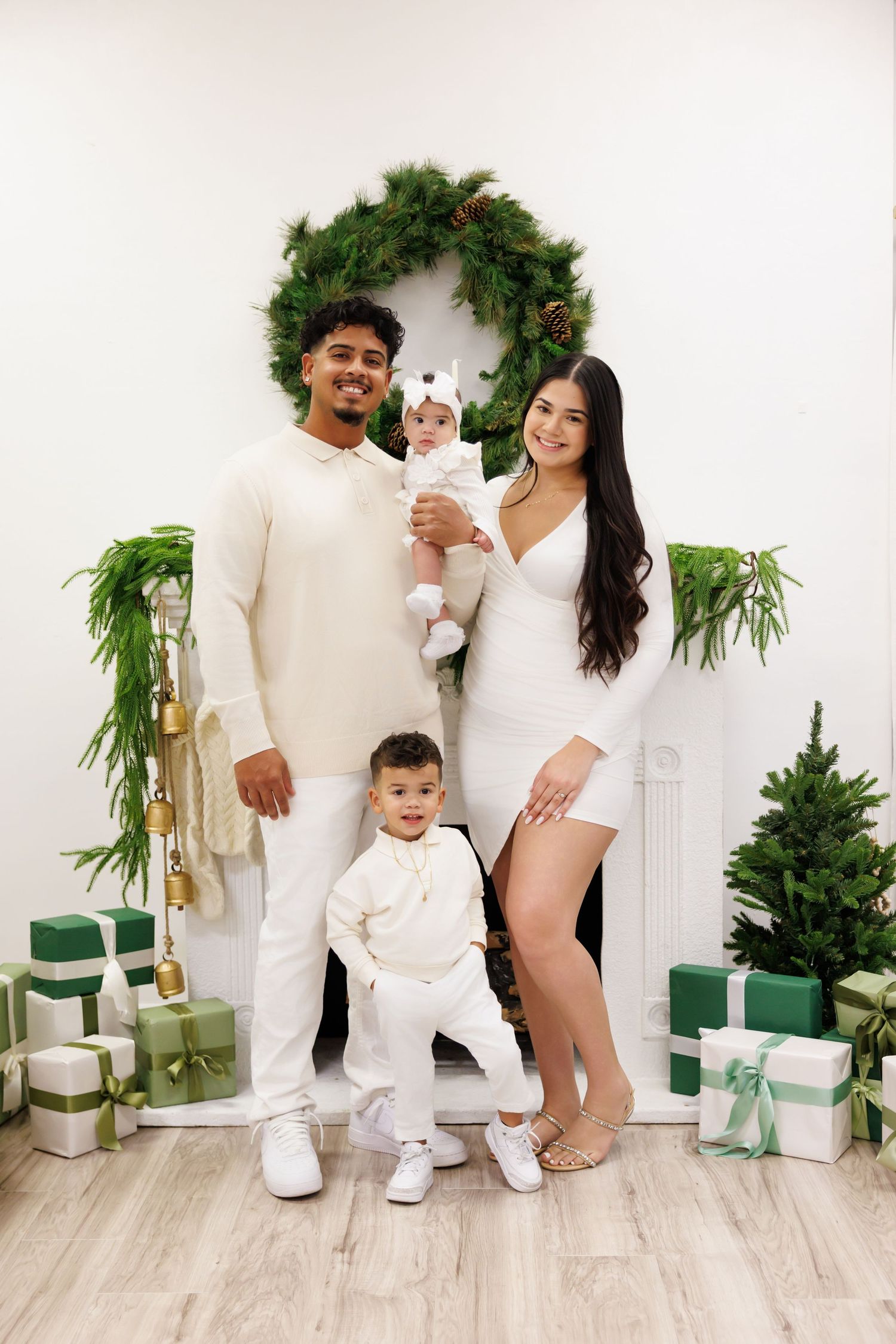 Family of four in matching white outfits posing by a decorated fireplace with Christmas wreaths and green gifts.