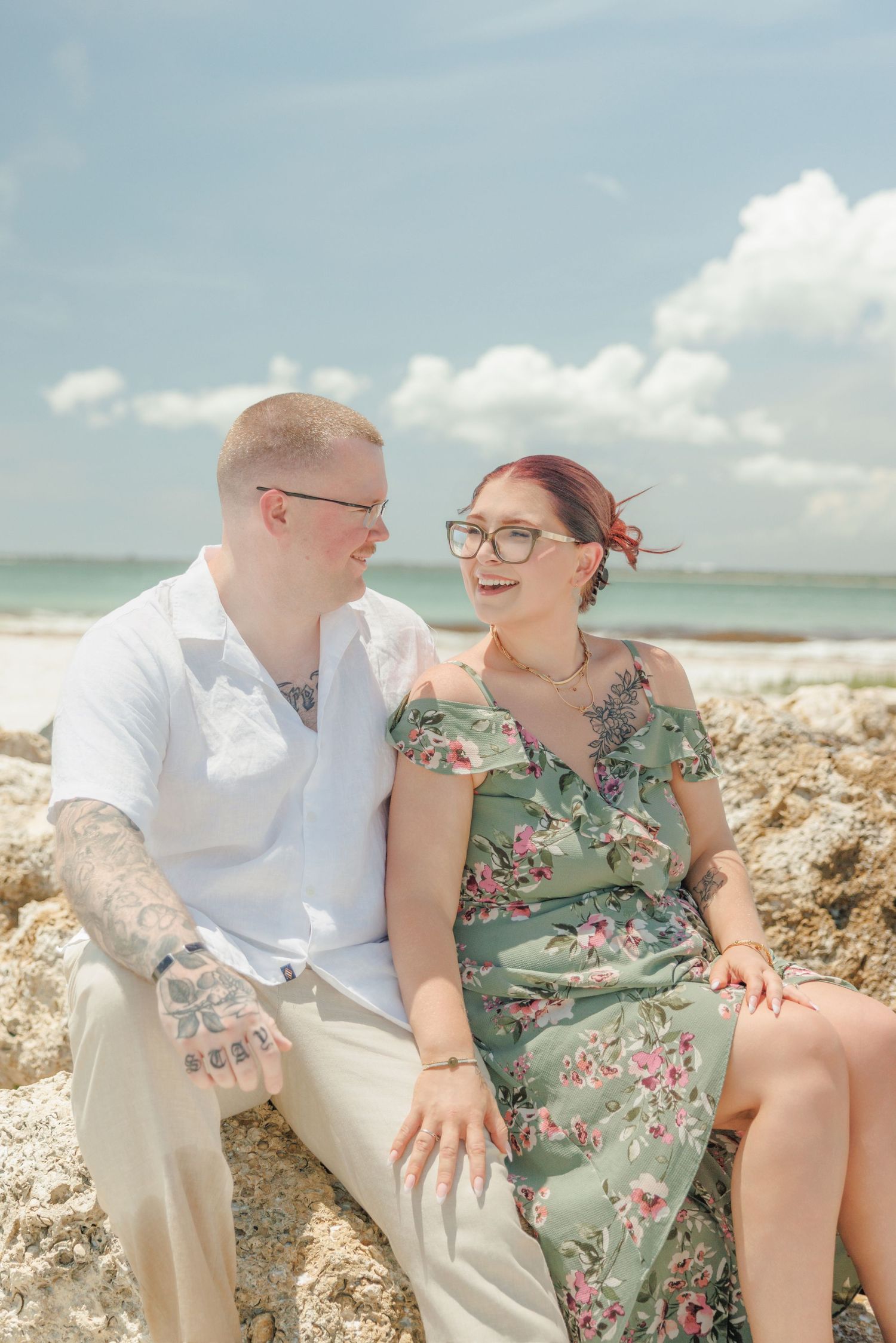 Smiling couple with tattoos and glasses sit close together on rocks at the beach, he in a white shirt and she in a floral green dress.