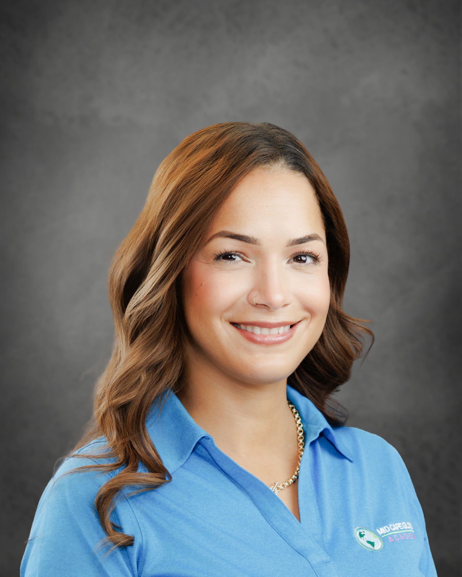 Smiling woman with long wavy auburn hair wearing a light blue collared polo shirt with a logo and a pearl necklace against a gray backdrop.