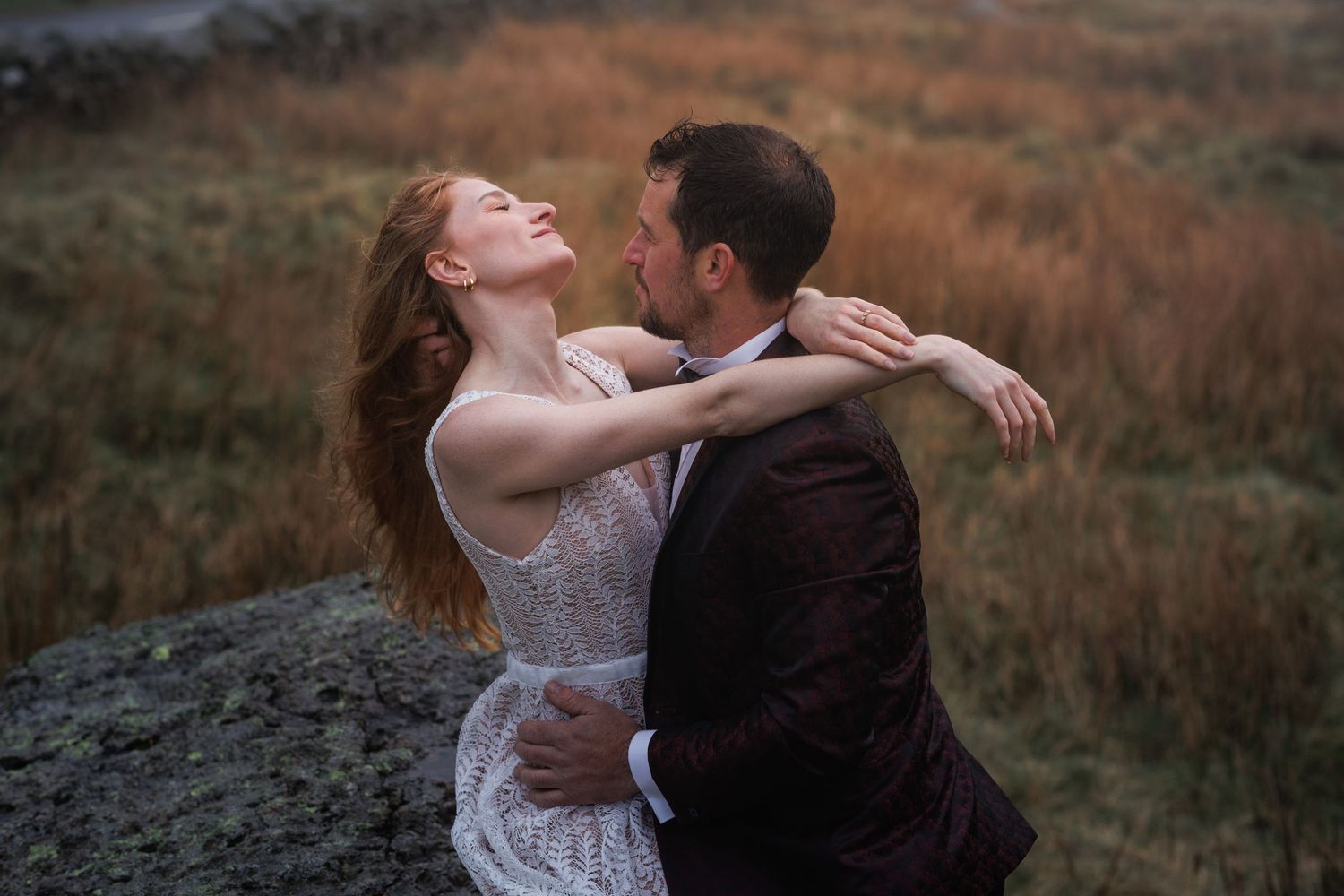 Two figures share an intimate embrace on rocky terrain at dusk with mountains in the background.