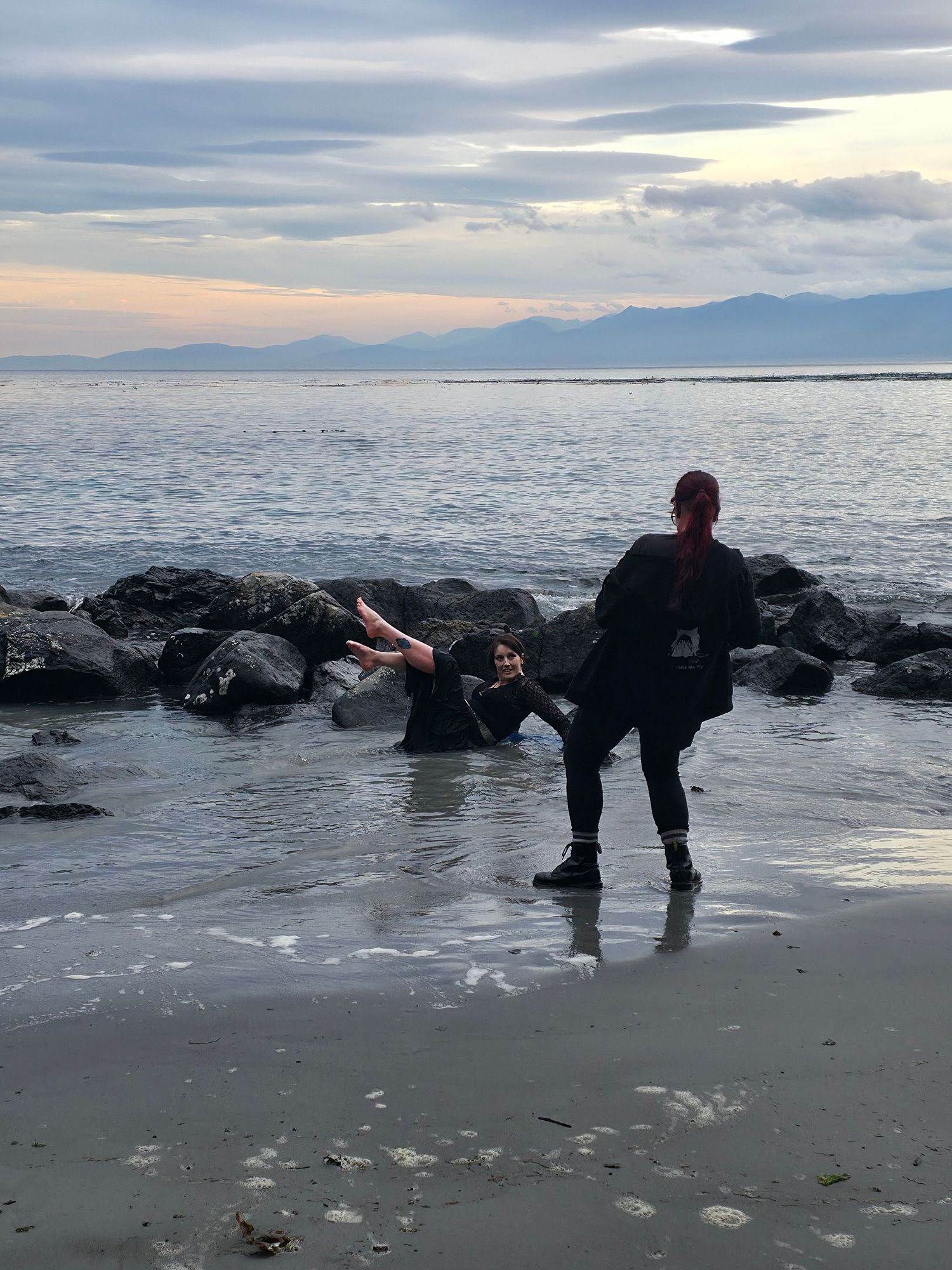A silhouetted figure performs a yoga pose on a rocky beach at sunset with mountains visible in the distance.