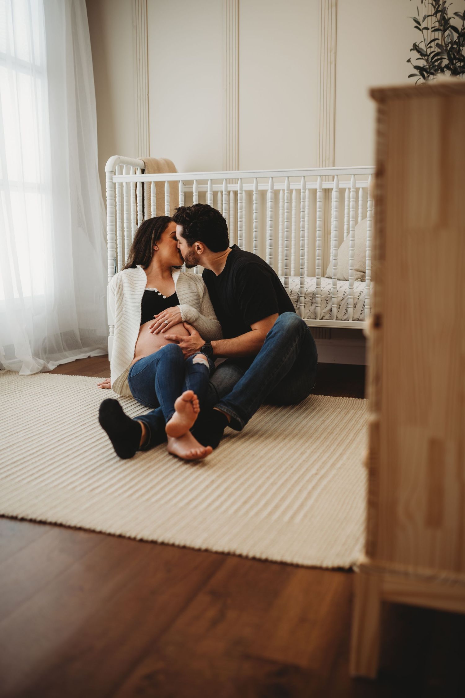 Couple sits together on carpet in nursery room with white crib and sheer curtains against natural lighting.