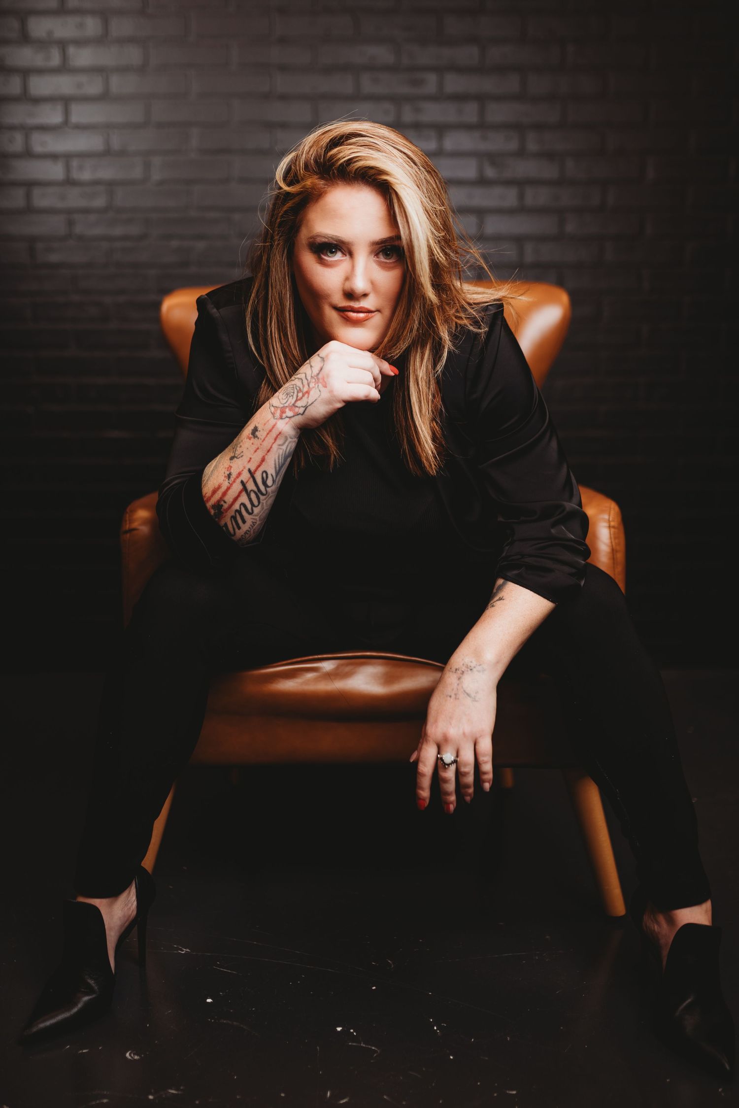 Portrait in a brown leather chair against dark brick wall featuring dramatic lighting and visible arm tattoos.