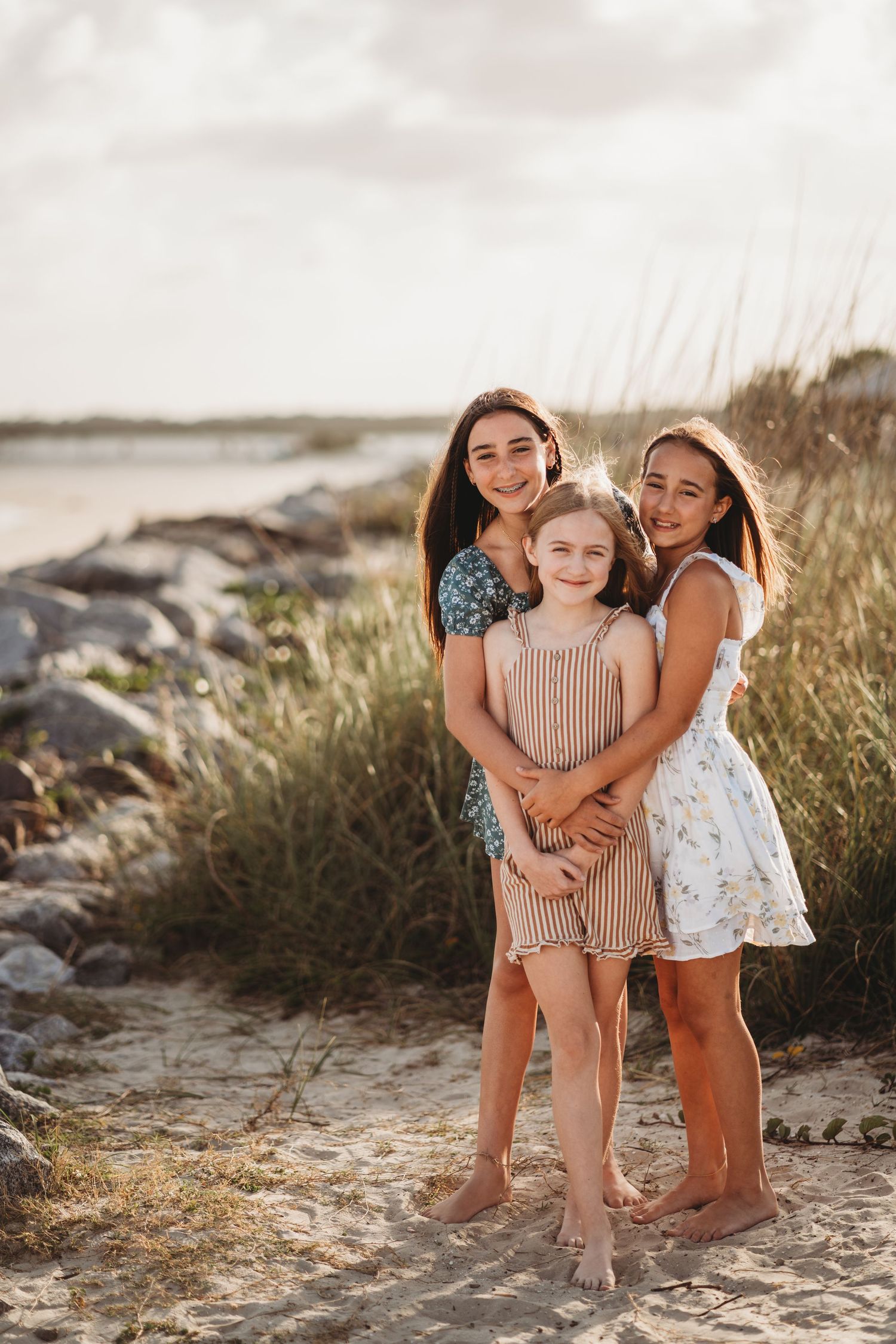 Three people embrace and smile while standing barefoot on a sandy beach at sunset with rocks and tall grasses nearby.