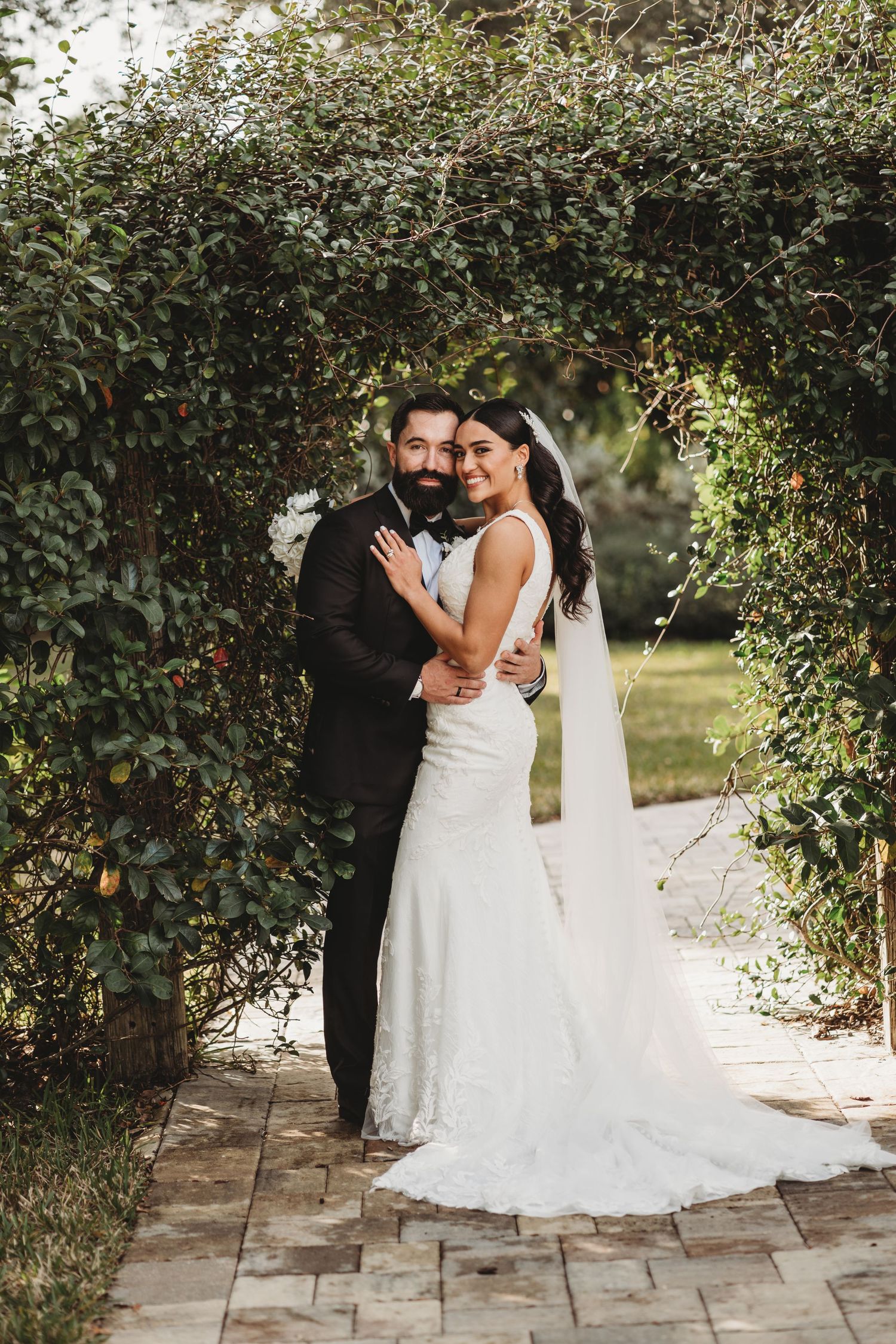 Newlyweds pose under a lush garden archway on a stone pathway.
